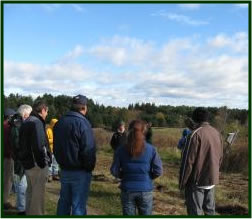 Group of people listening to a person speaking on the edge of a field with forest in the distant background.