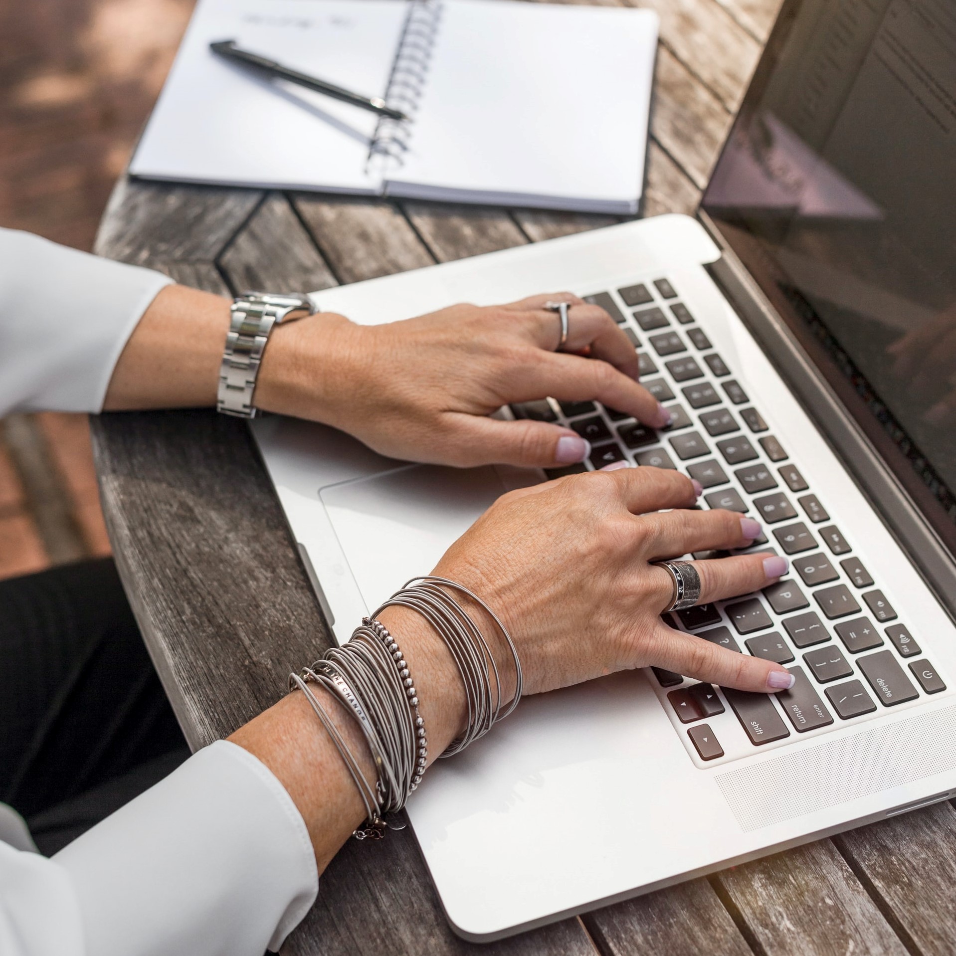 hands with bracelets and rings hovering over laptop keyboard next to pad of paper and pencil