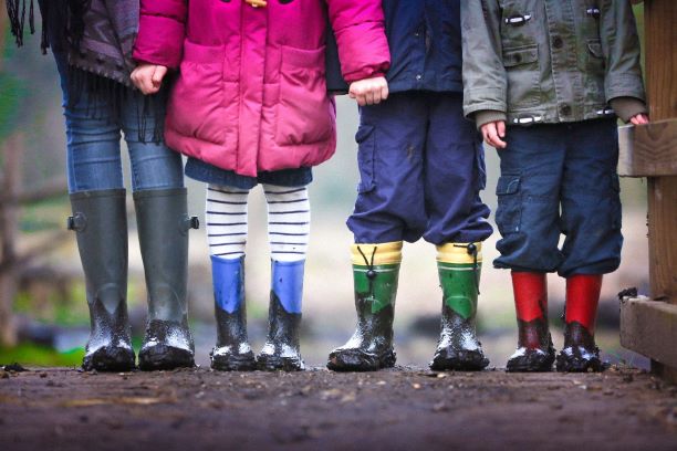 4 young children standing side by side outside, wearing rain boots