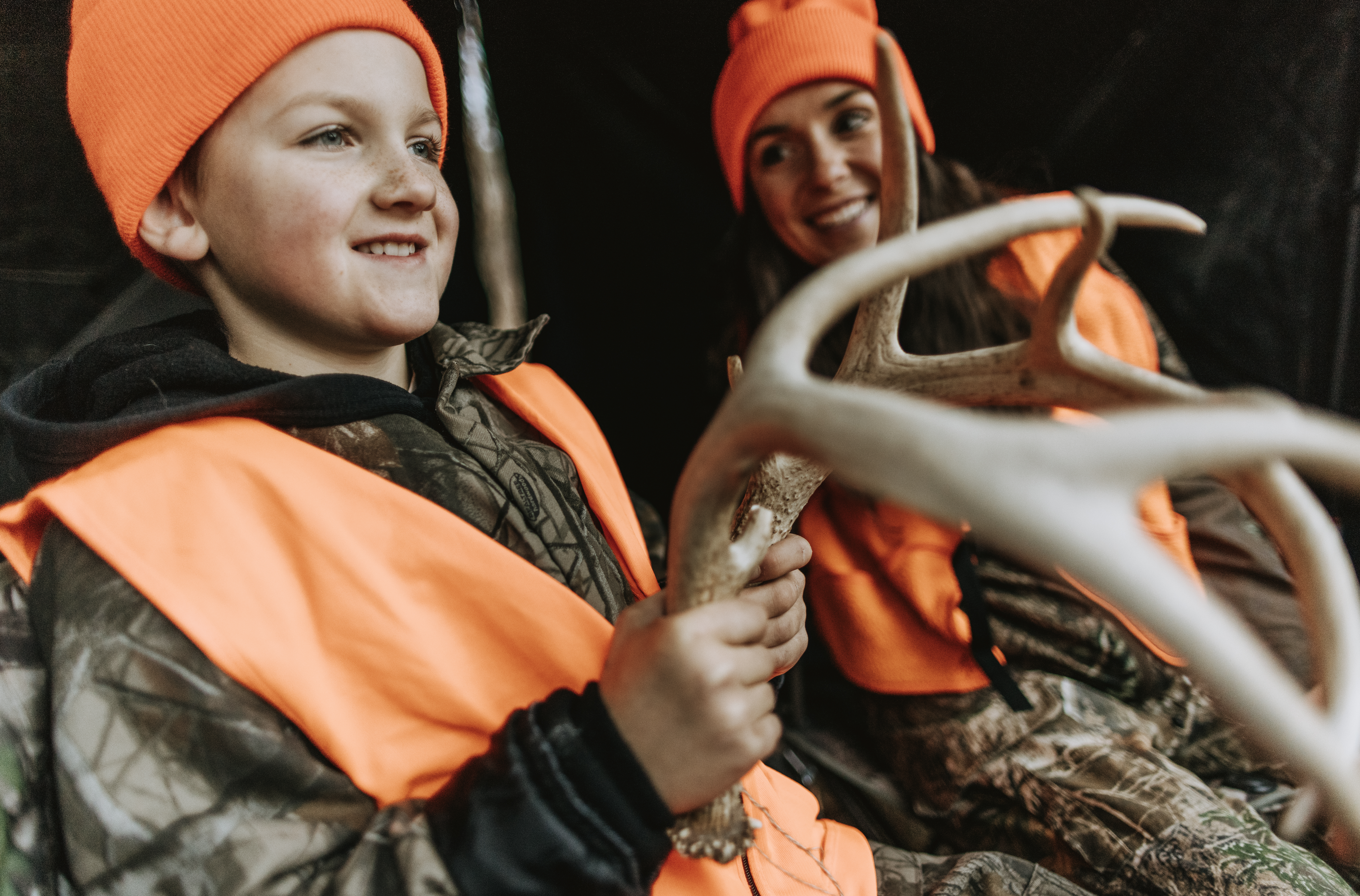A mother and son in a hunting blind, smiling and holding deer antlers.