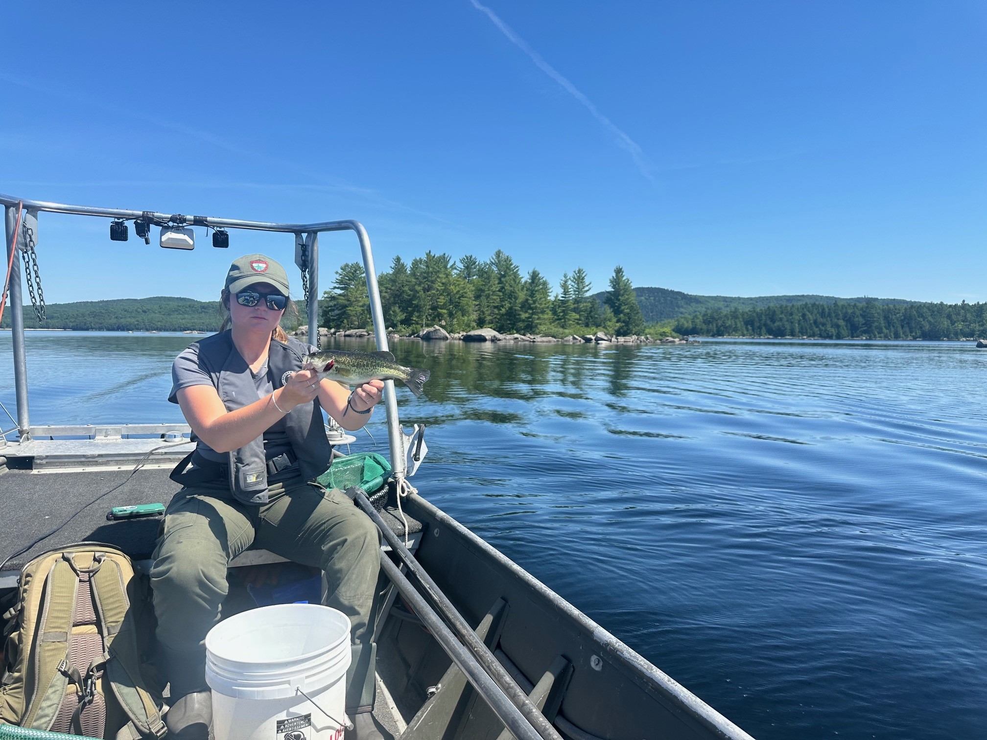 fisheries biologist holding largemouth bass with sad expression and beautiful mountain and lake background