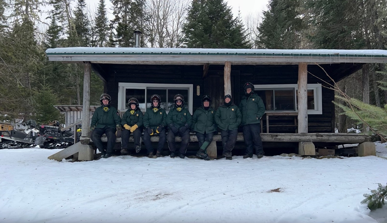 Group of game wardens in snowmobile gear standing in front of a remote warden camp with lots of snow