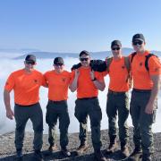 Five game wardens wearing blaze orange tshirts and green pants at the top of a mountain with blue skies