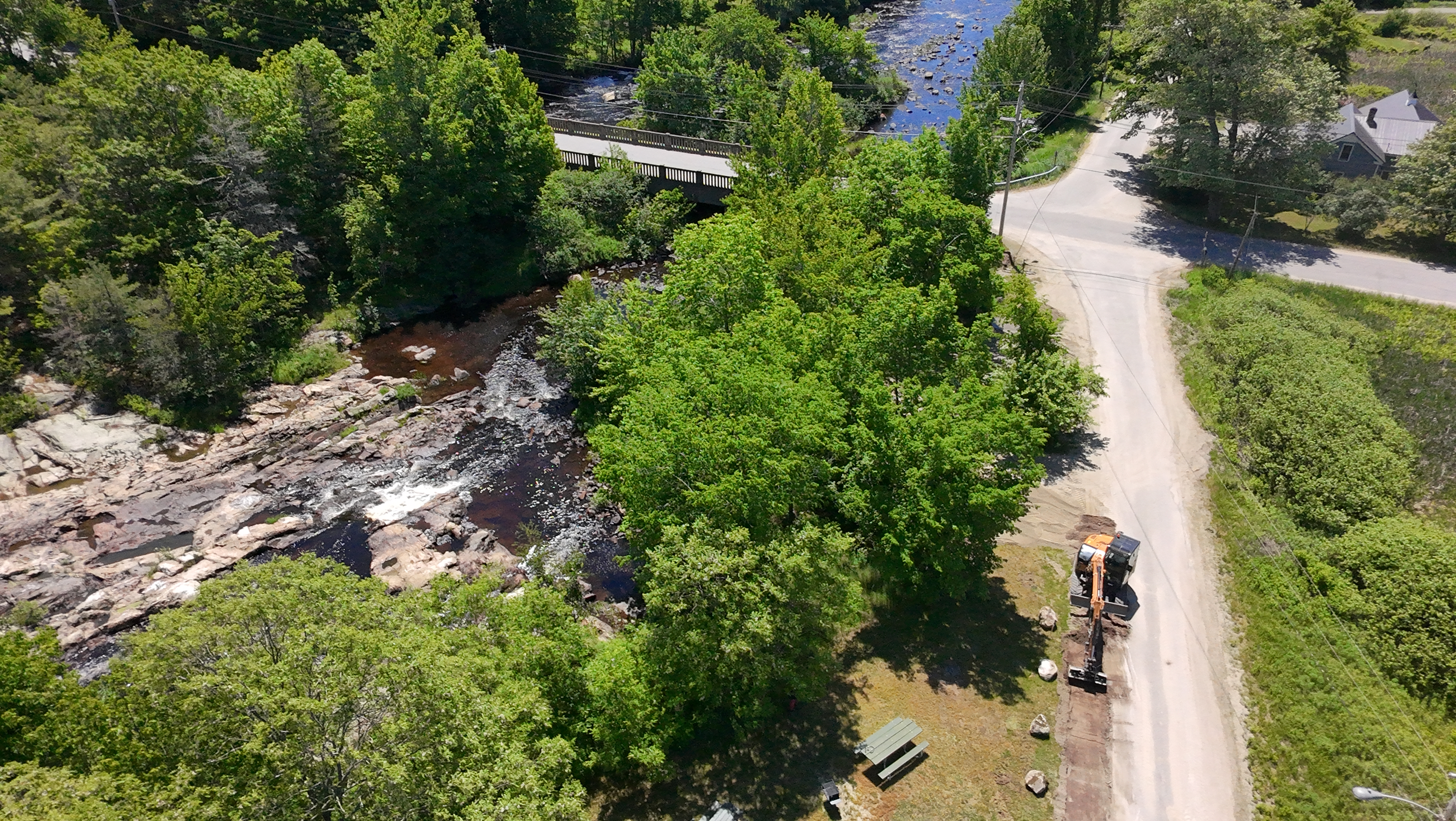 Aerial view of an excavator surrounded by a river, trees, and a dirt road.
