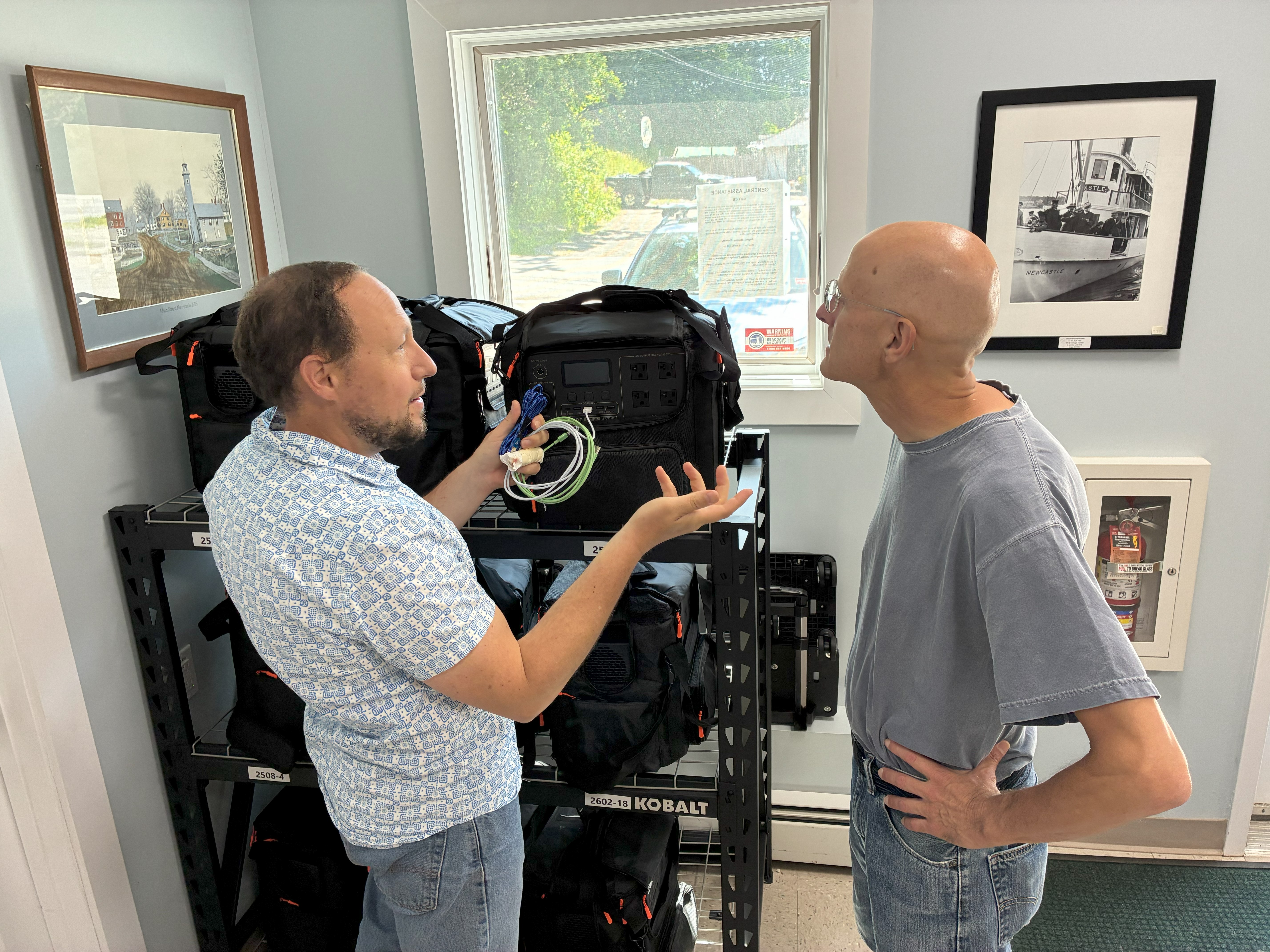 Two men stand next to a rack of black bags in a town office.