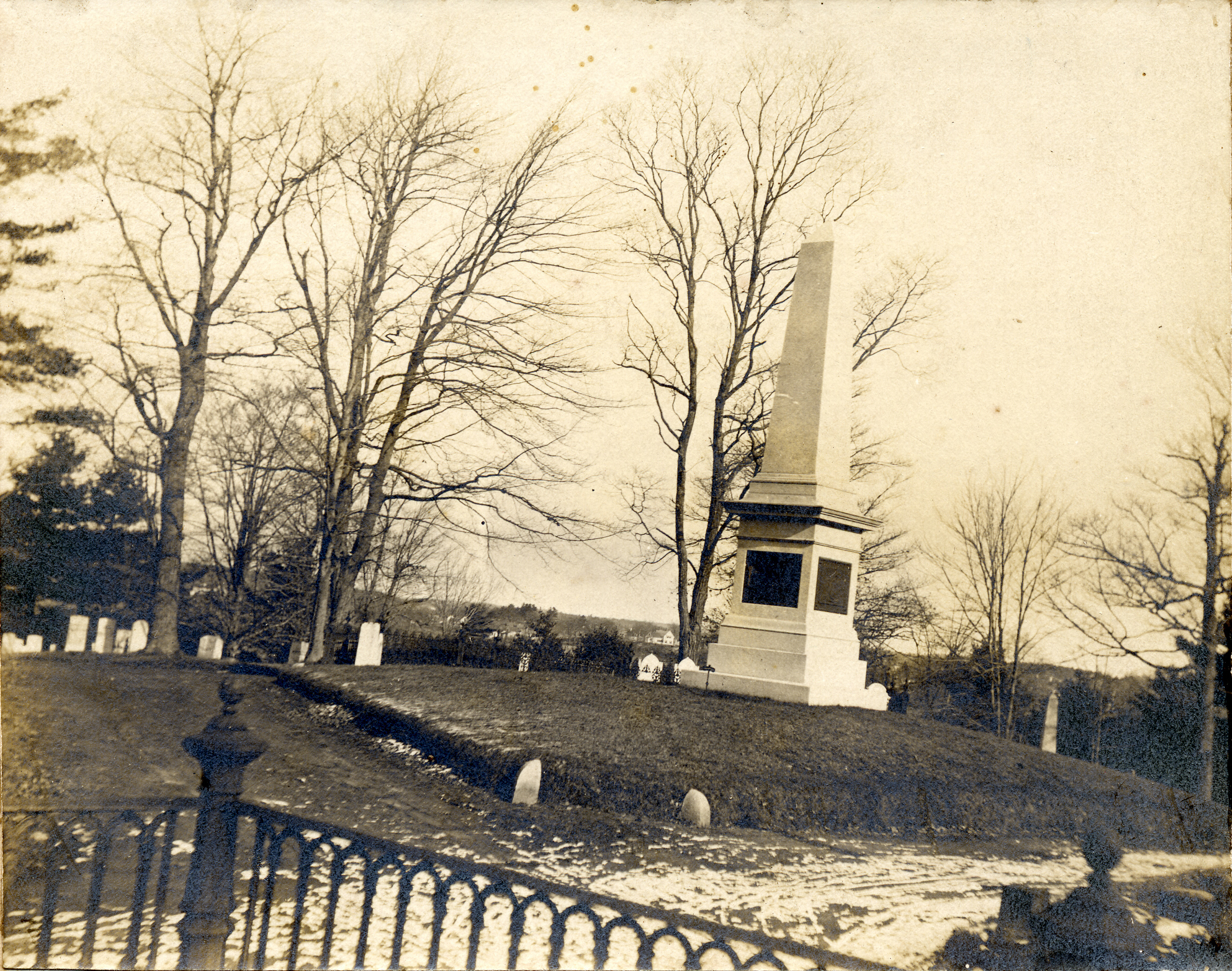 A vintage sepia-toned photograph of a tall stone obelisk monument standing on a grassy hill within a cemetery, surrounded by bare trees and smaller headstones.