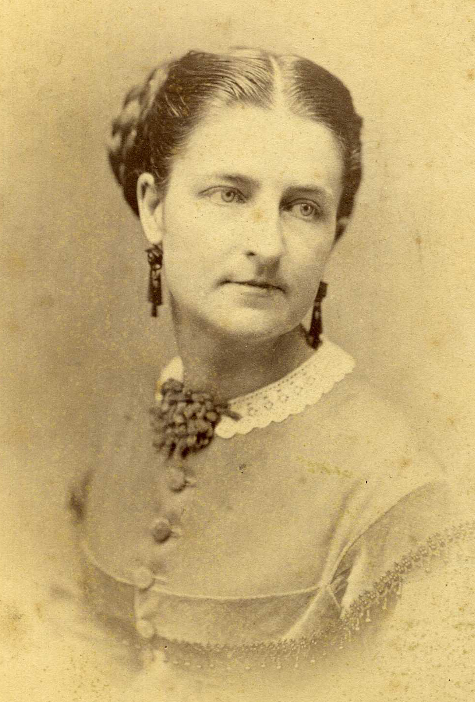 A vintage sepia-toned bust portrait of a woman with her hair styled up, facing slightly right. She is wearing dark, dangling earrings and a light-colored high-collared dress adorned with a lace neckpiece.