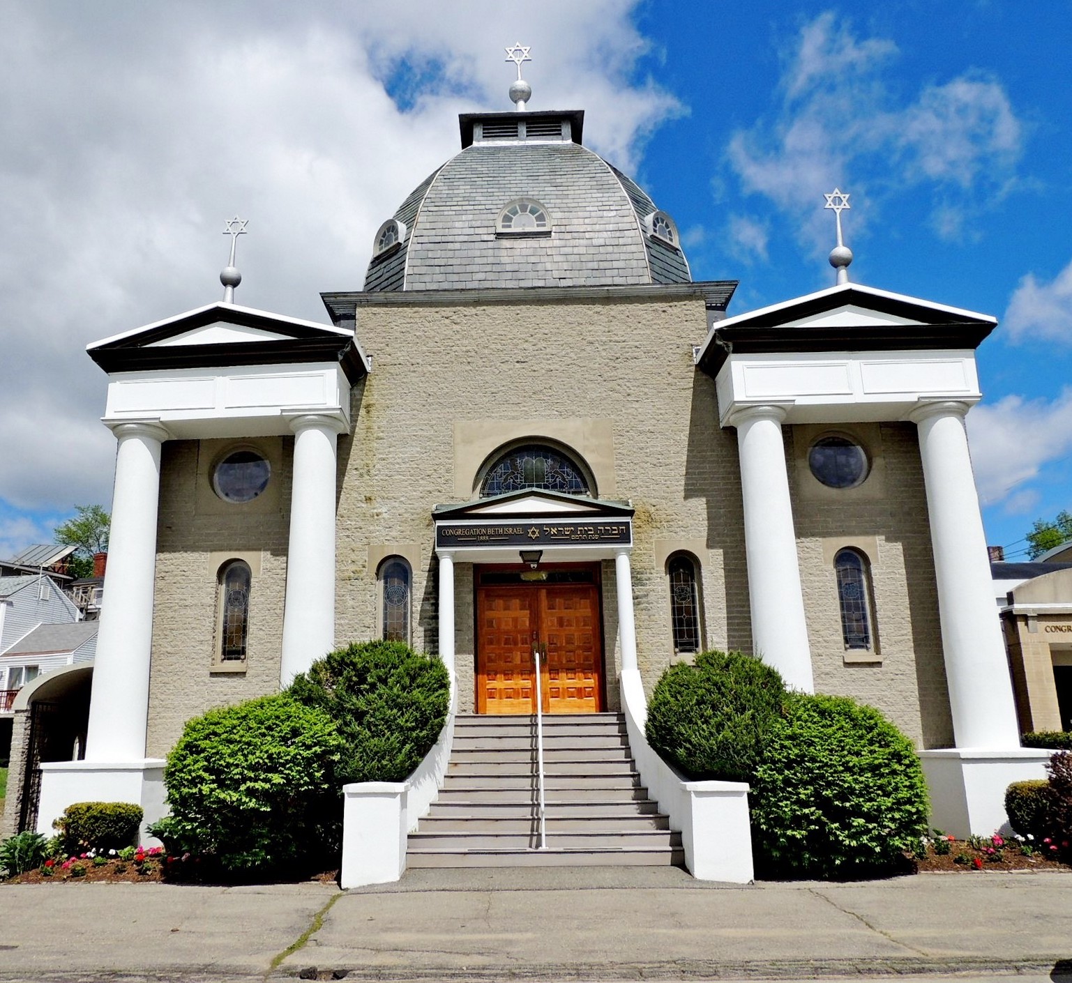 A historic synagogue featuring light brick walls, a central grey dome, and white columns flanking the front facade. Steps lead up to double wooden doors topped by a sign reading "CONGREGATION BETH ISRAEL" and Hebrew text, with silver Stars of David serving as roof finials against a blue sky.