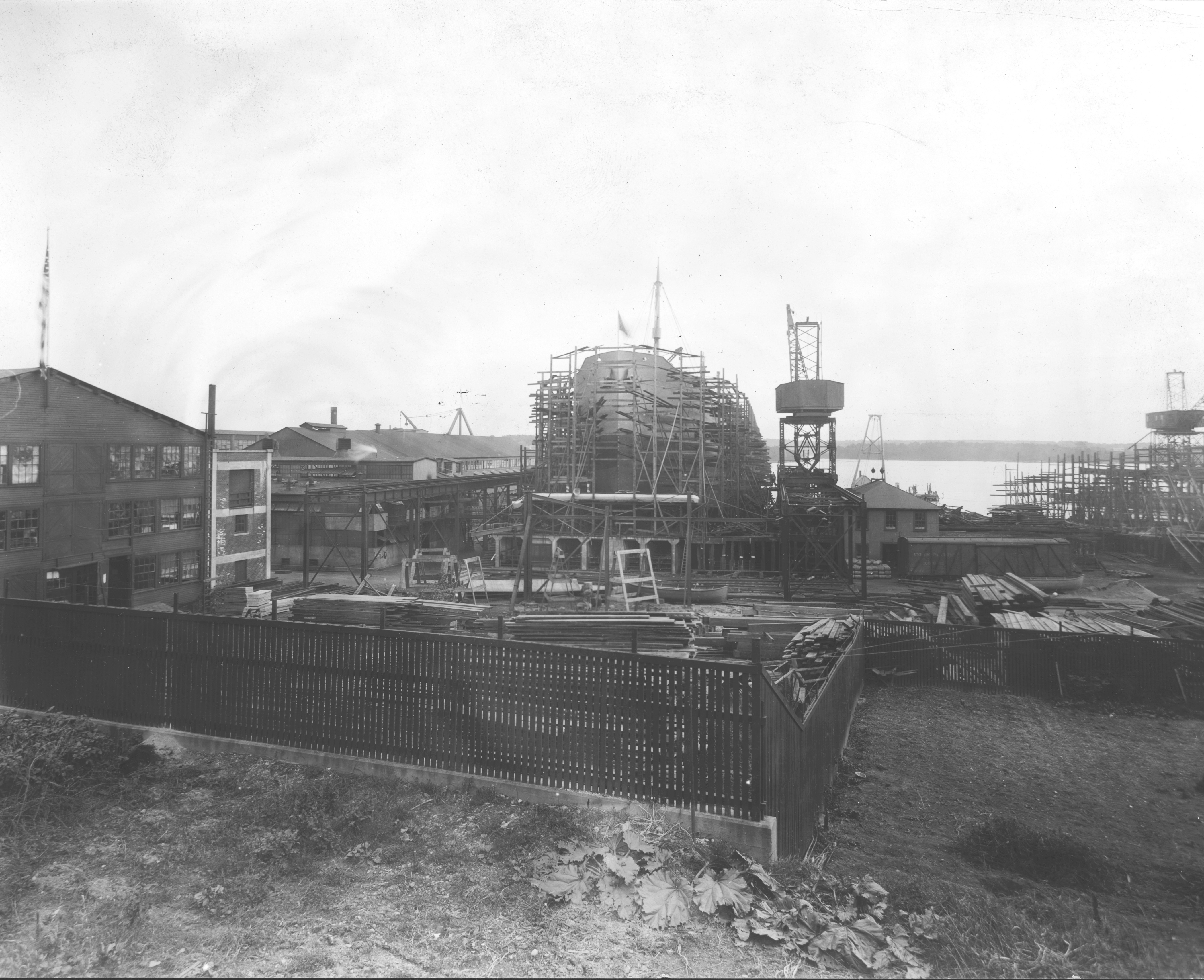 A black and white historical photograph showing a wide view of a busy shipyard with industrial buildings, cranes, and a large ship hull under construction inside a scaffolding framework.