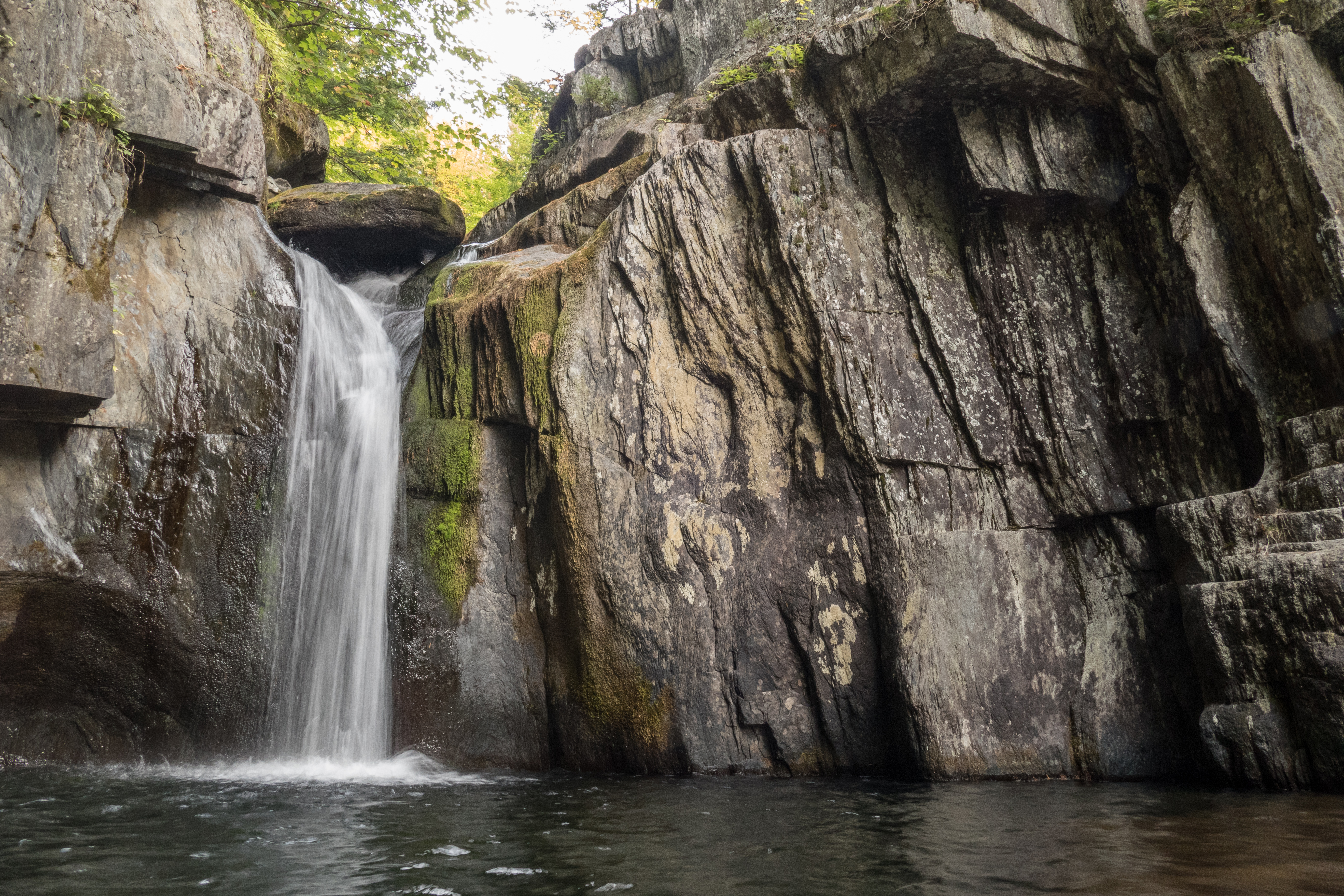 A close-up shot of a waterfall pouring over a rugged, layered rock wall into a dark pool below. Moss grows along the damp crevices of the steep, gray rock face.