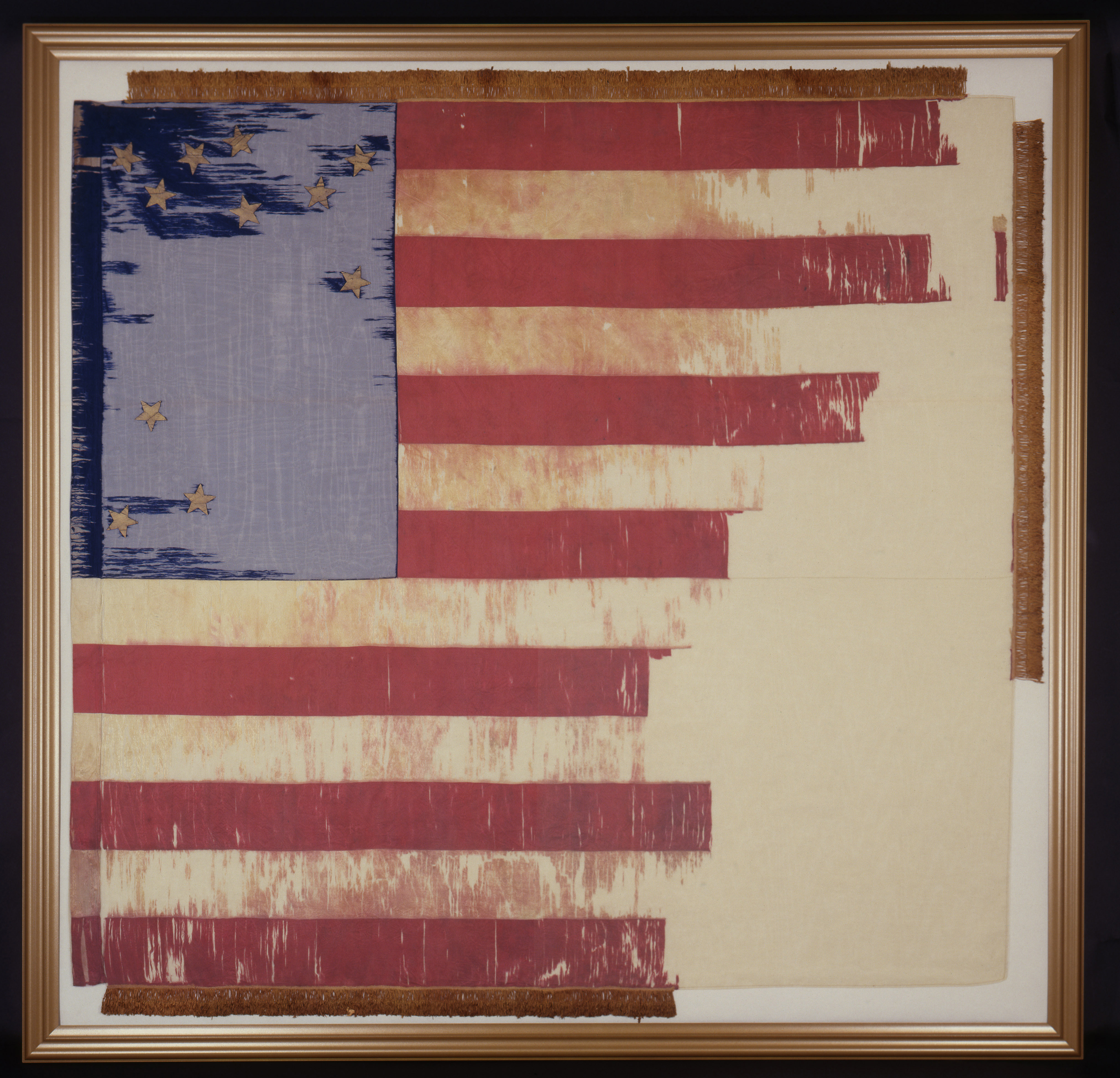 A heavily worn and frayed historic American flag displayed inside a square gold frame. The flag features faded red and white stripes with a blue canton in the top left corner bearing gold stars arranged in a circular pattern, with a few stars separated at the bottom and right.