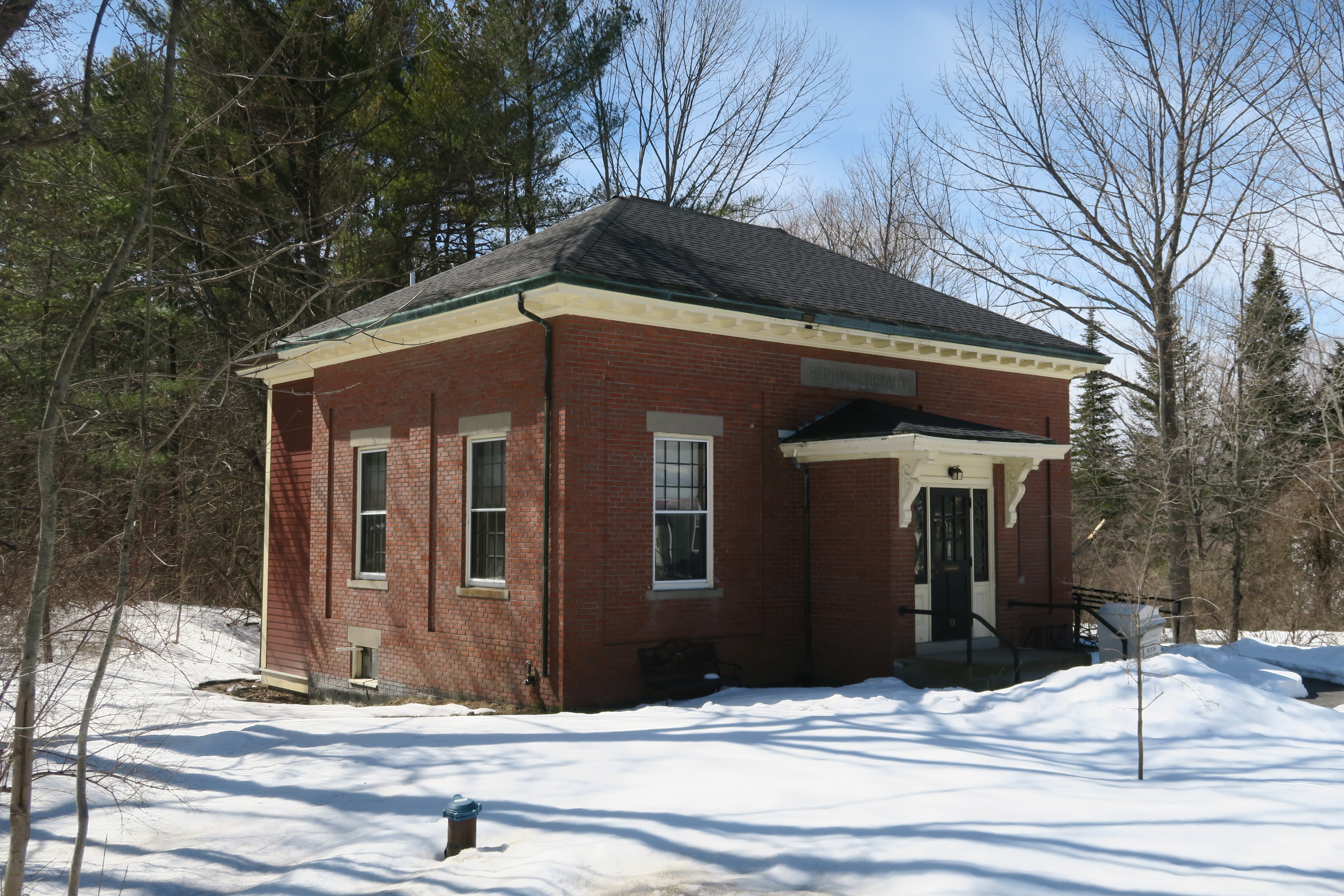 A single-story brick library building with a dark hipped roof and a small covered entrance porch, surrounded by patches of snow on the ground and bare trees under a blue sky.