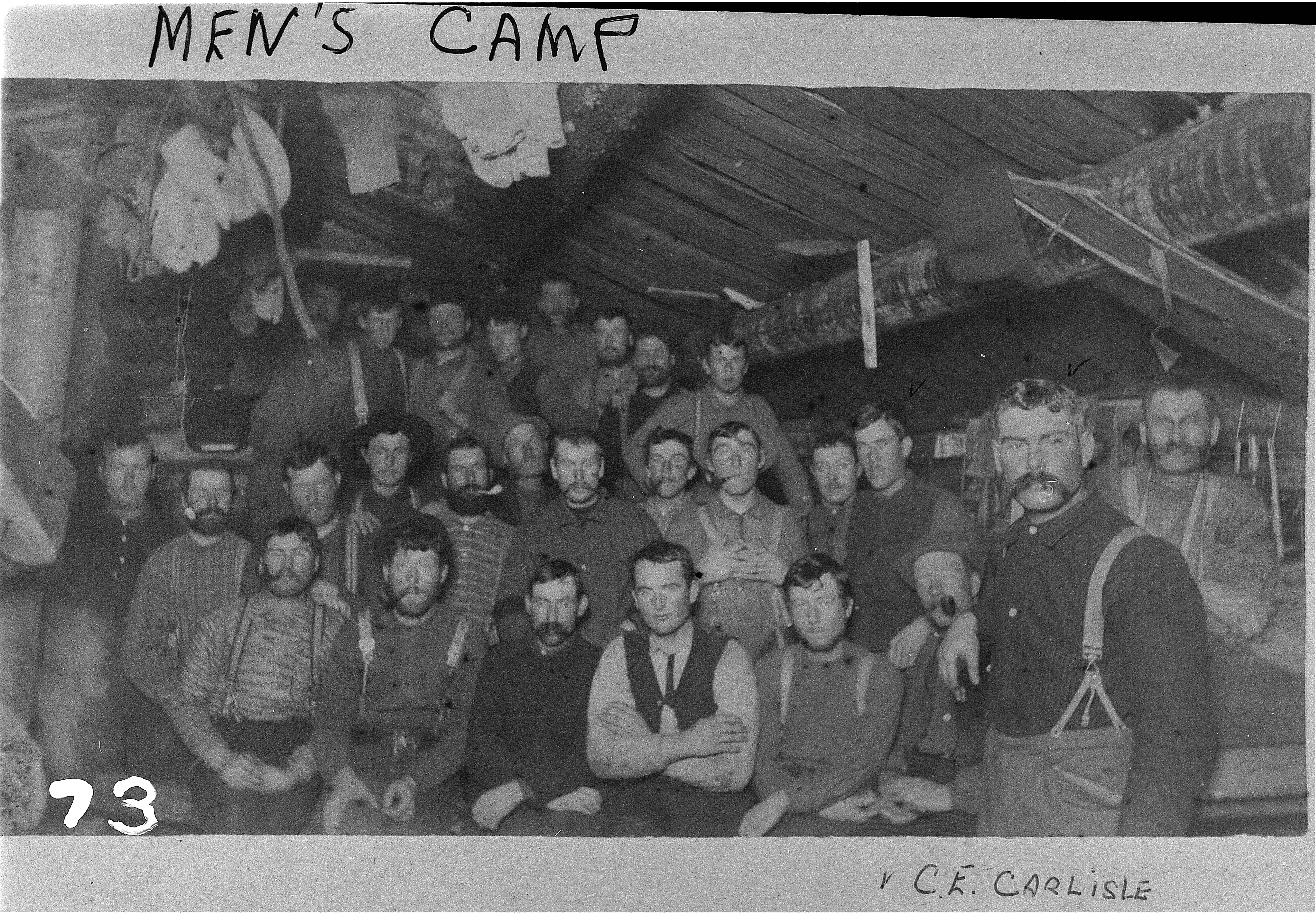 A vintage black and white group photo inside a rustic, low-ceilinged log cabin bunkhouse. A large group of male loggers, many sporting thick mustaches and suspenders, sit tightly packed together looking toward the camera.