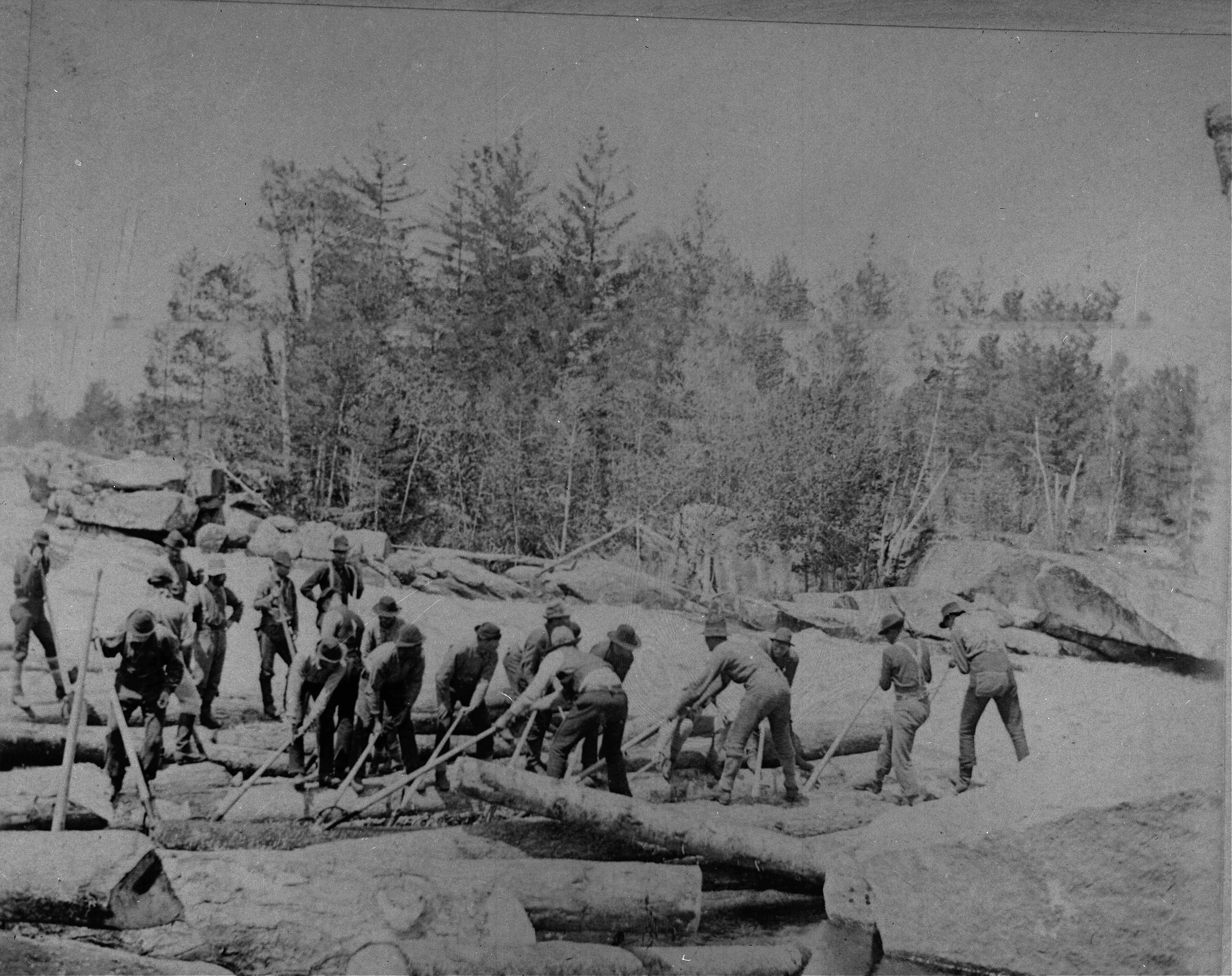 A historic black and white photograph of a large crew of loggers working along a rocky riverbank. The men are using long wooden poles to maneuver logs through the shallow water against a backdrop of pine trees.