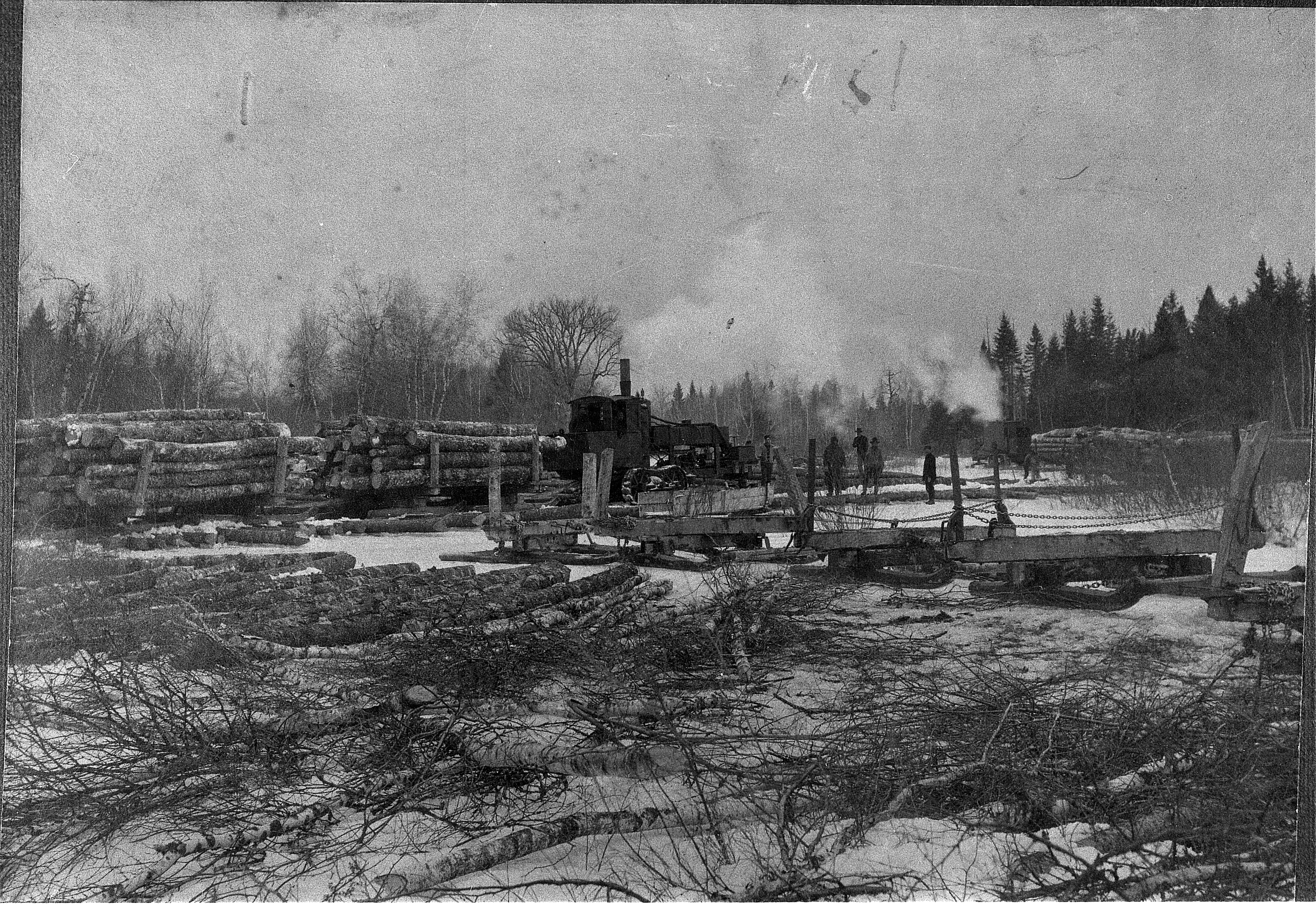 A black and white winter photograph showing an early steam-powered log hauler on tracks positioned in a clearing. Several logging sleds loaded with timber sit nearby on the snow-covered ground surrounded by a sparse forest.