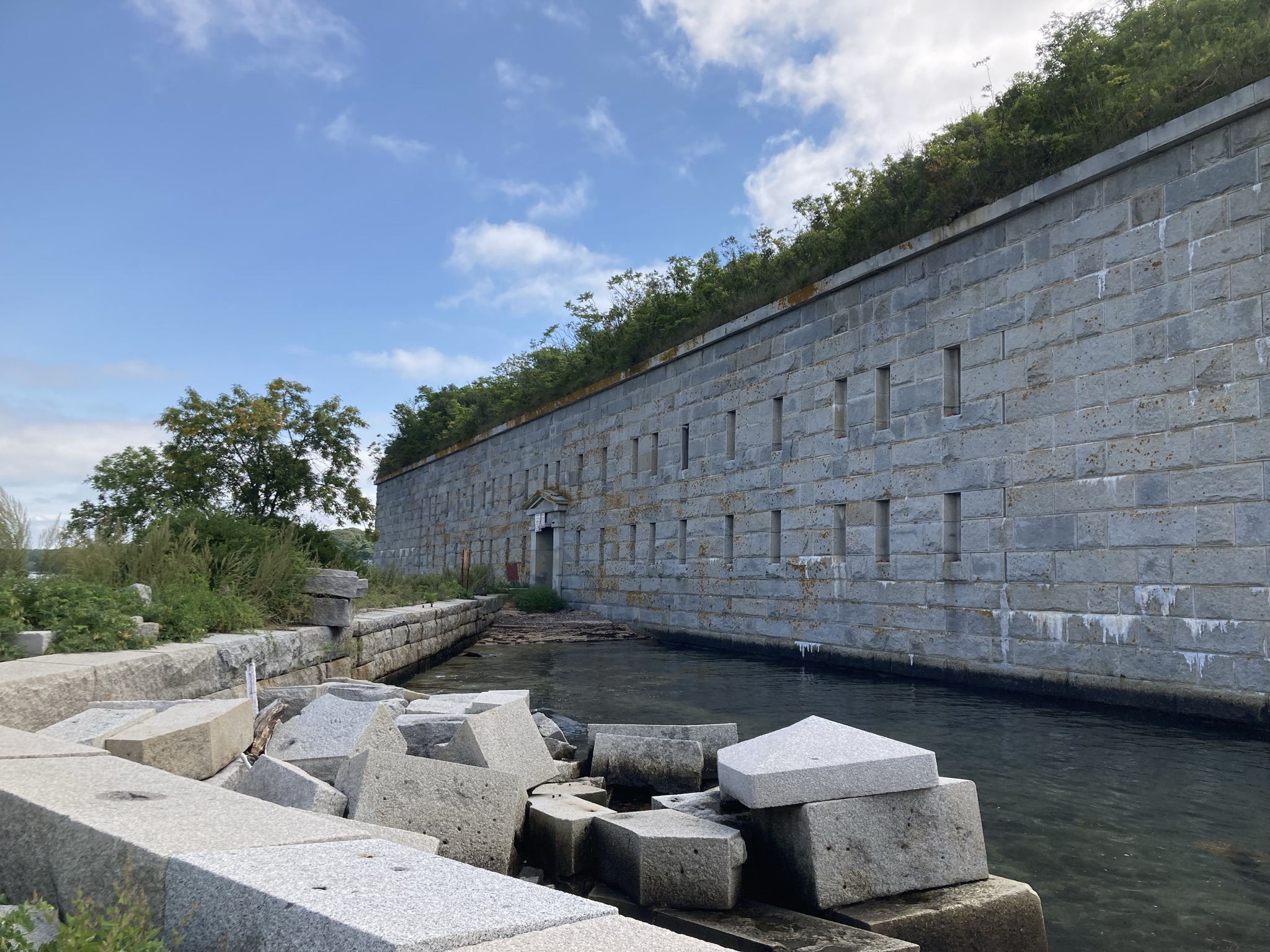 A massive granite stone fortress wall with narrow window slits curves along a calm body of water under a blue sky, with large loose blocks of stone piled in the foreground.