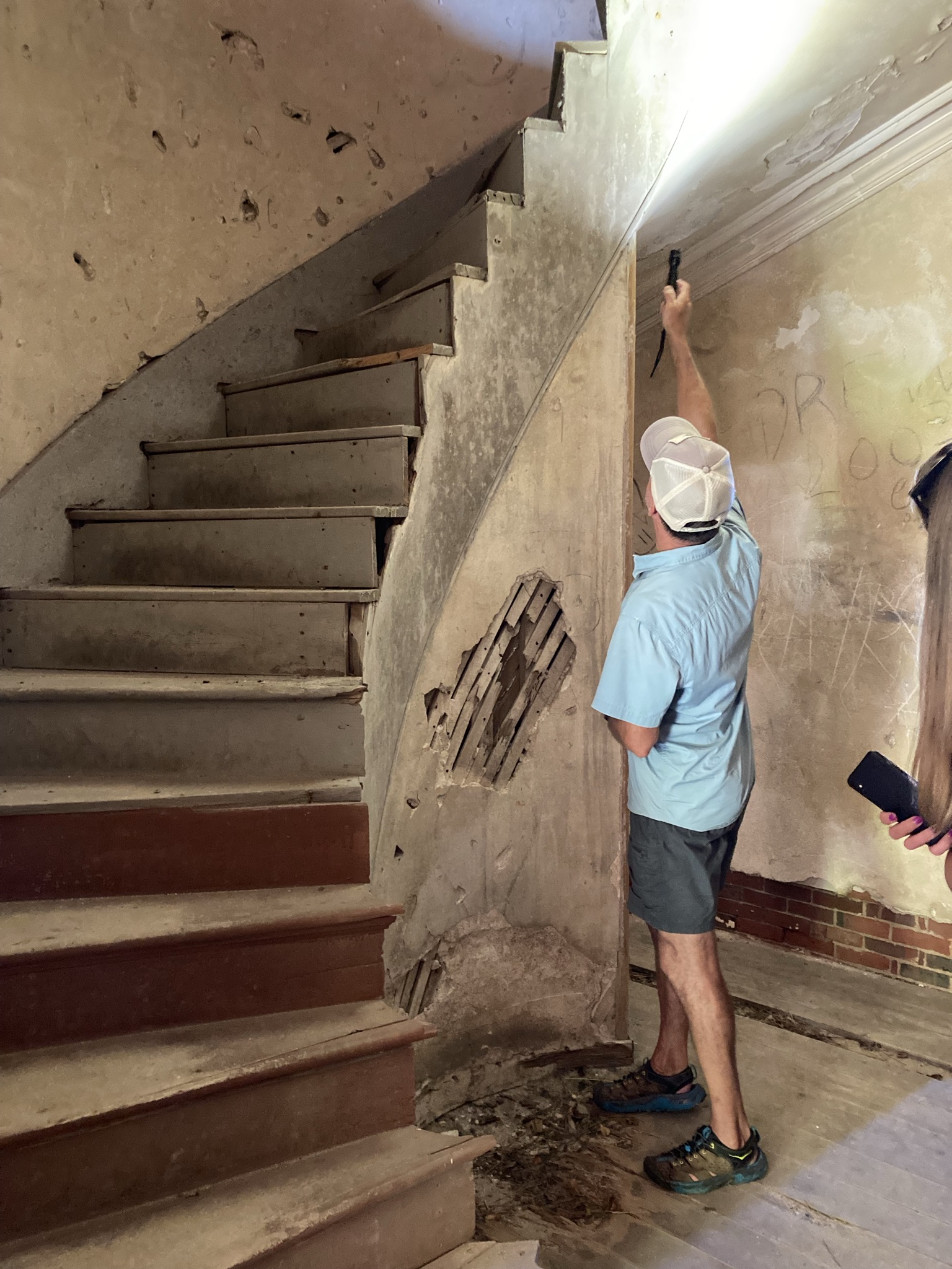 Inside a historic building, a man wearing a baseball cap and blue shirt uses a flashlight to inspect a crumbling wall near an old wooden staircase.