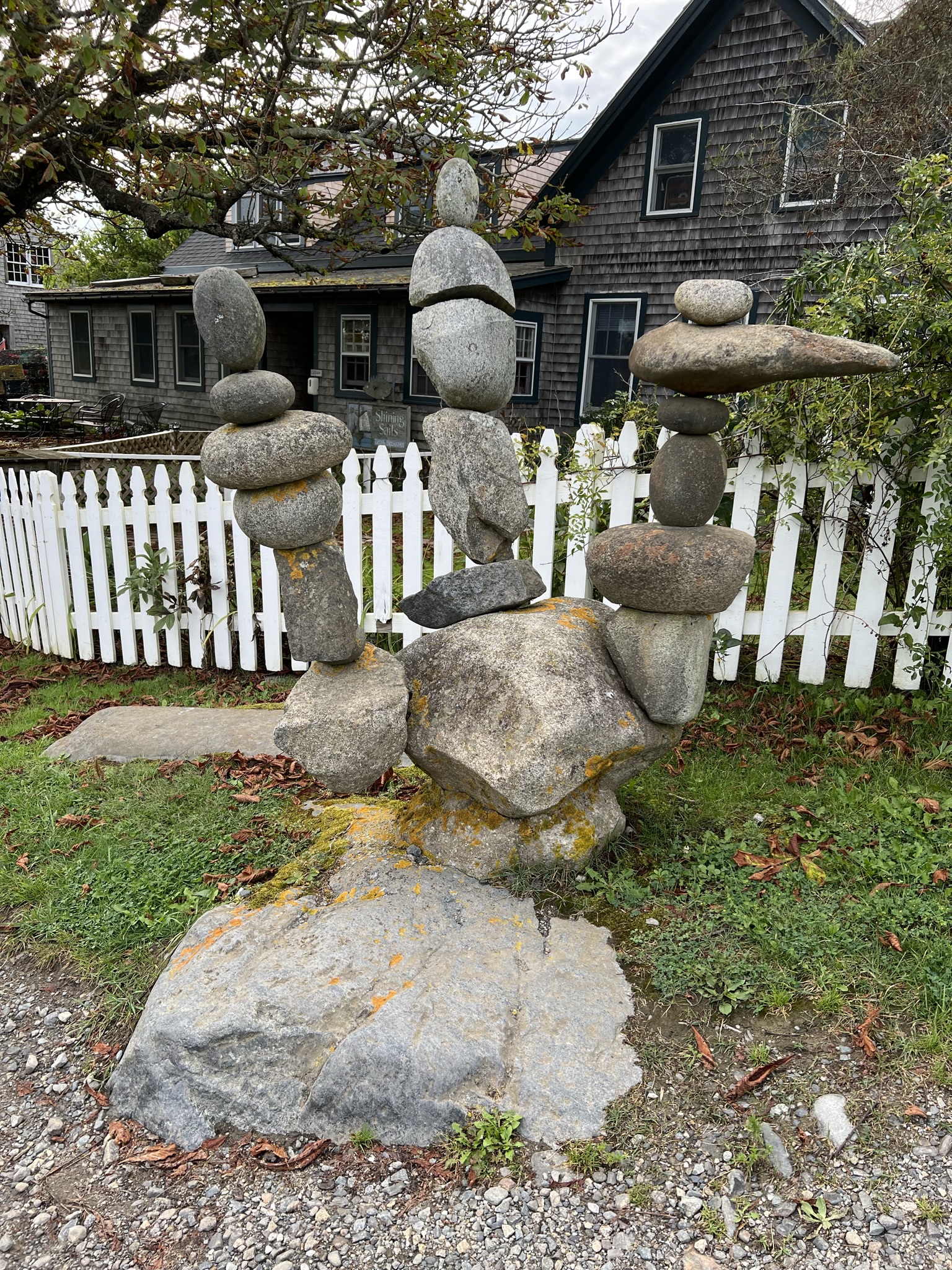 A close-up photograph of a unique rock sculpture made from several balanced, stacked gray stones forming a shape that resembles a hand gesturing a peace sign or three vertical fingers. The sculpture sits on a mossy boulder in front of a white picket fence and a shingled house.