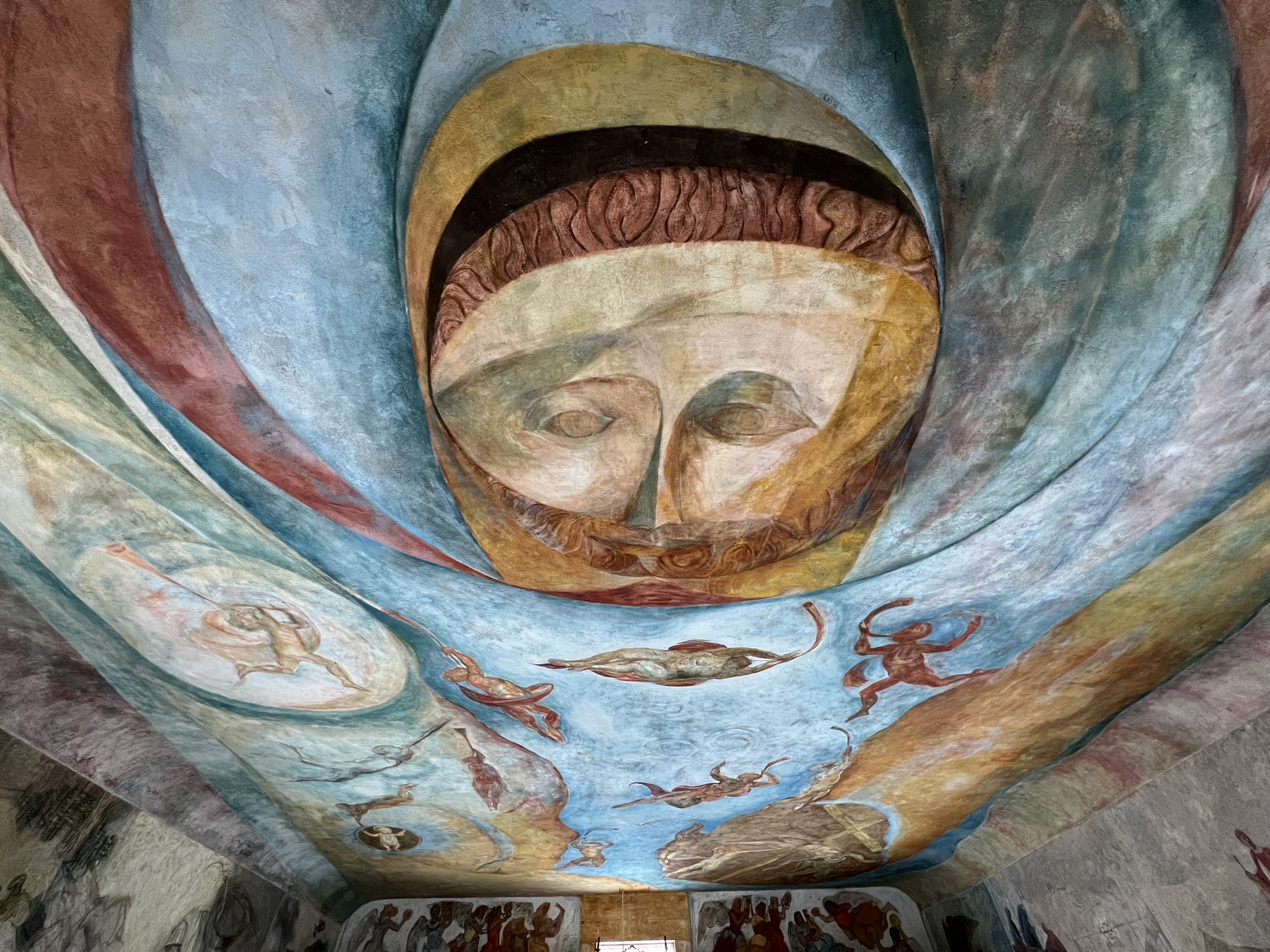 Close-up of the meetinghouse ceiling mural, dominated by a large painted face with closed eyes at center, surrounded by swirling blue and gold tones and small figures in motion.