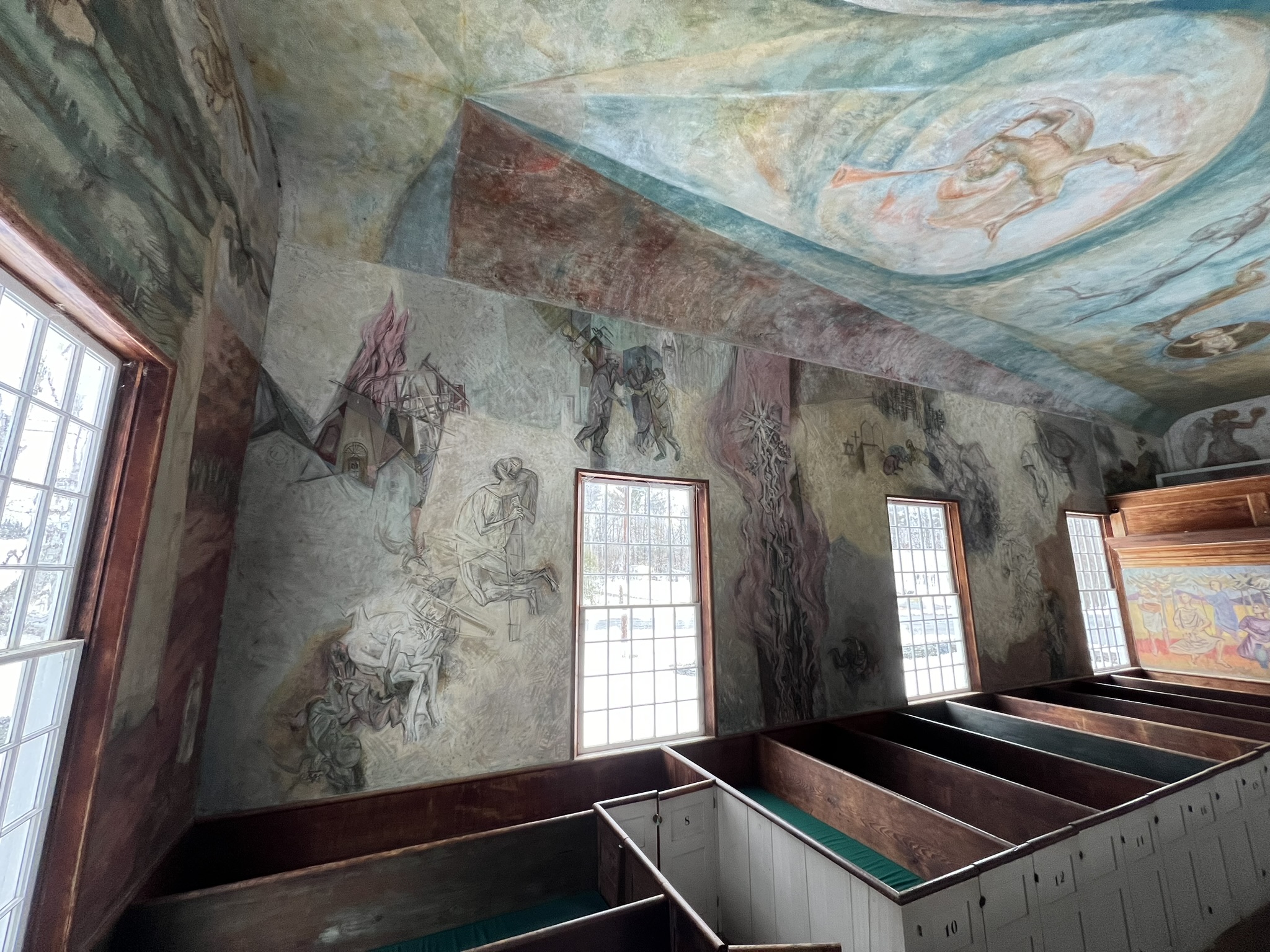 Interior side-wall view of the meetinghouse showing partially completed mural sketches depicting figures, flames, and symbolic imagery in muted grays and pinks, with box pews below and a dynamic ceiling mural above.