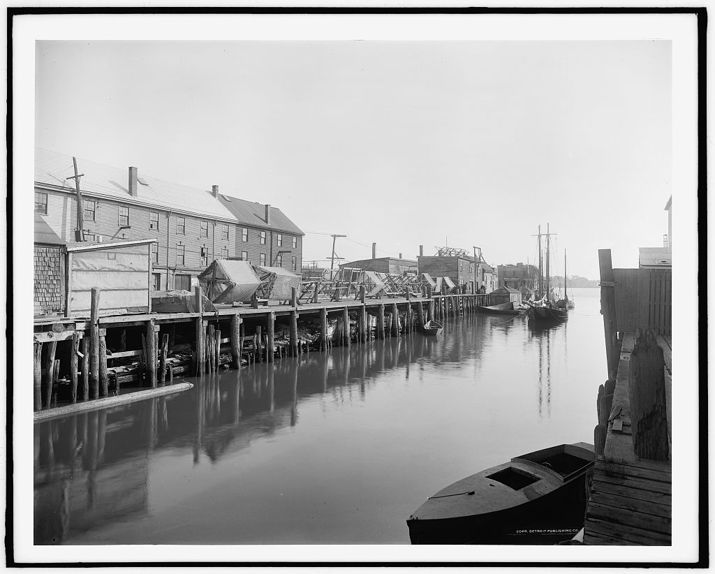 A vintage black and white photograph looking down a narrow harbor waterway between wooden piers in Portland, Maine. A long wooden dock supported by numerous pilings extends along the left side, stacked with large, hexagonal wooden frames used for drying fishermen's nets next to a row of multistory wooden waterfront buildings. A small boat rests in the foreground, and a larger masted vessel is moored in the distance.