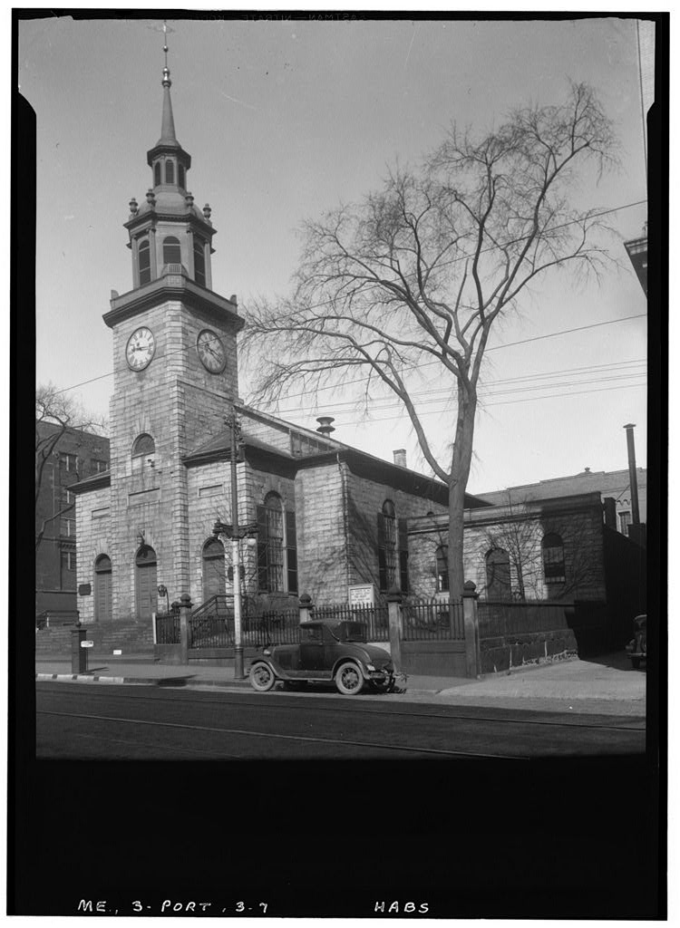 A black and white architectural photograph of a historic stone church featuring a prominent clock tower and tiered steeple, with a vintage automobile parked on the street in front of its fenced-in yard.