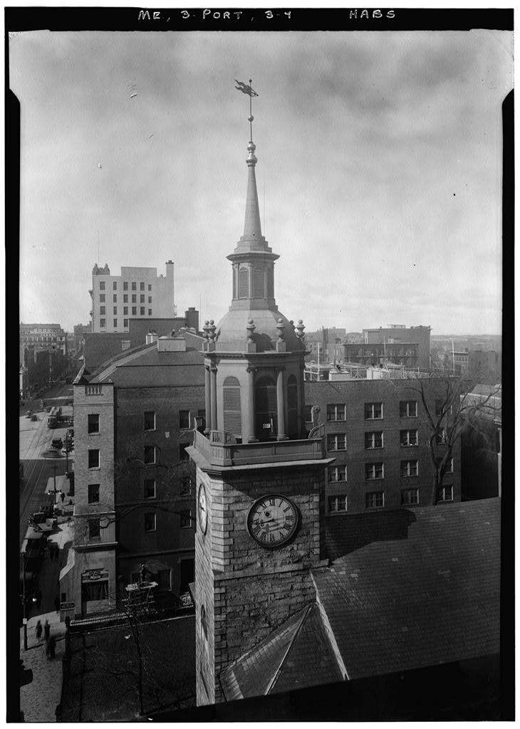 An elevated black and white photograph capturing a close-up perspective of a stone church's clock tower and weather-vaned steeple, overlooking surrounding multi-story brick city buildings and a street below.