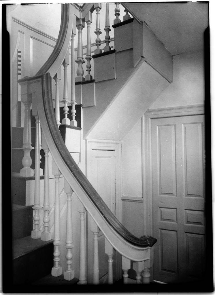 Black-and-white archival photo of the Tate House entrance hall staircase, featuring a gracefully curved handrail, turned balusters, and paneled wainscoting on the adjacent wall.