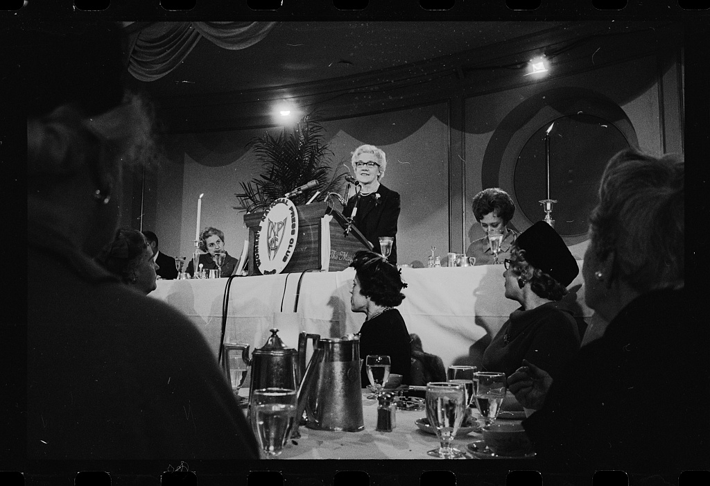 A woman speaking at a podium during a formal banquet, with attendees seated at tables.