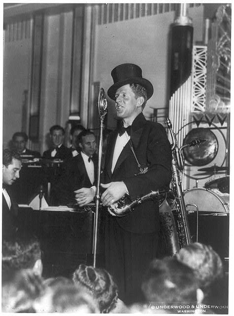 A black and white photograph of a man in a tuxedo and top hat standing on a stage at a microphone, holding a saxophone, with an orchestra visible in the background.
