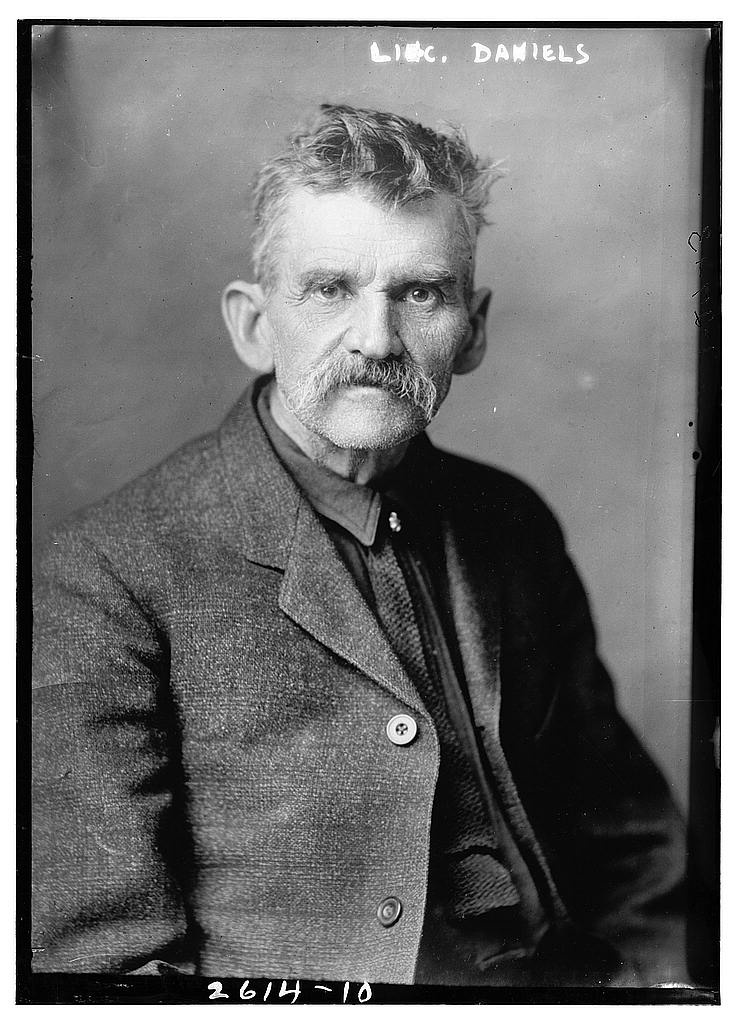 A black and white close-up photographic portrait of an older man with tousled gray hair, a full mustache, and an intense expression, wearing a dark shirt and a textured jacket. Handwritten text at the top reads "LINC. DANIELS."