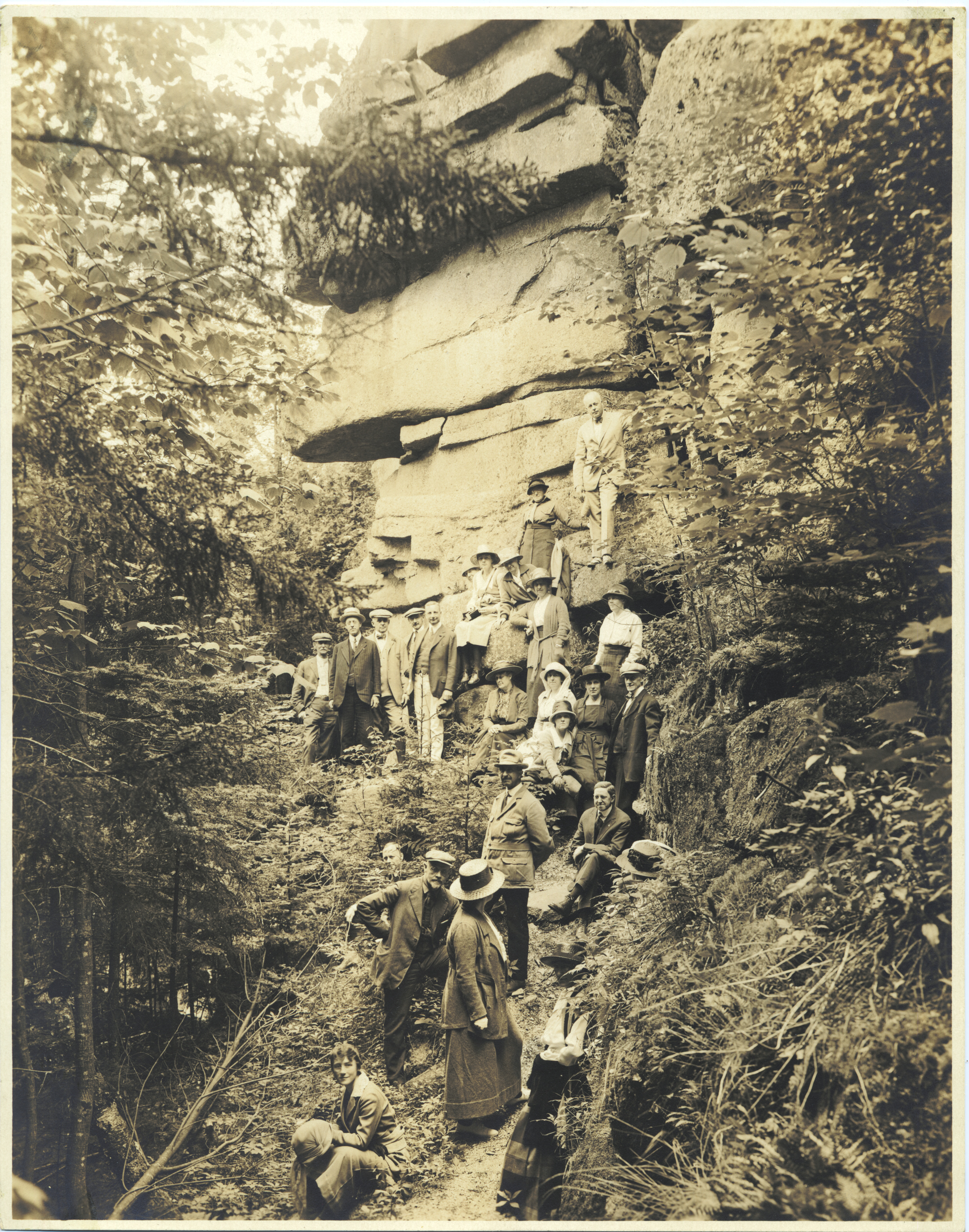 A vintage sepia photograph of a large group of early 20th-century tourists in formal attire posing along a rugged, forested trail beneath a massive granite cliff formation.