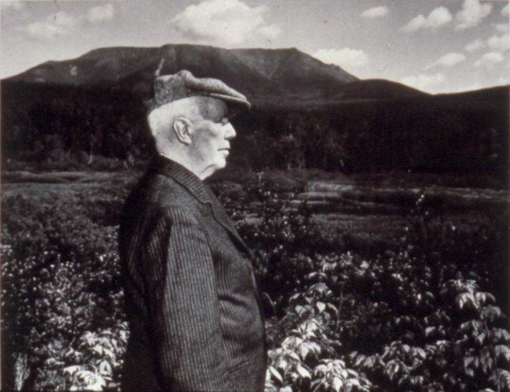 A historic black and white photograph in profile of an elderly man wearing a flat cap and a striped suit jacket, looking out over a dense field toward Mount Katahdin in the background.