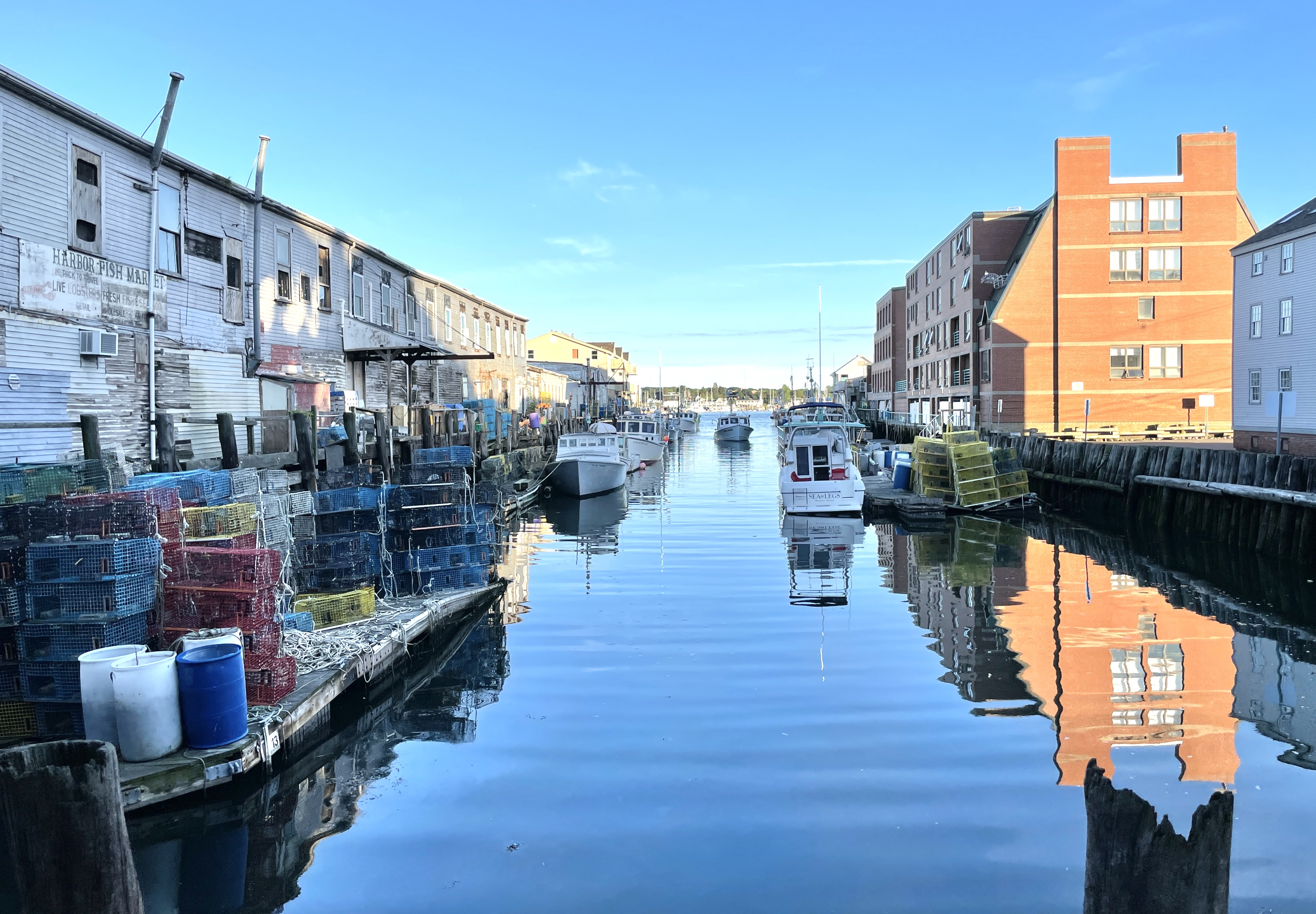 A daytime view looking down a narrow harbor slip flanked by buildings. On the left, stacks of colorful lobster traps sit on a floating dock next to weathered wooden structures, while the right side features a modern brick building reflecting in the still blue water where several boats are moored.