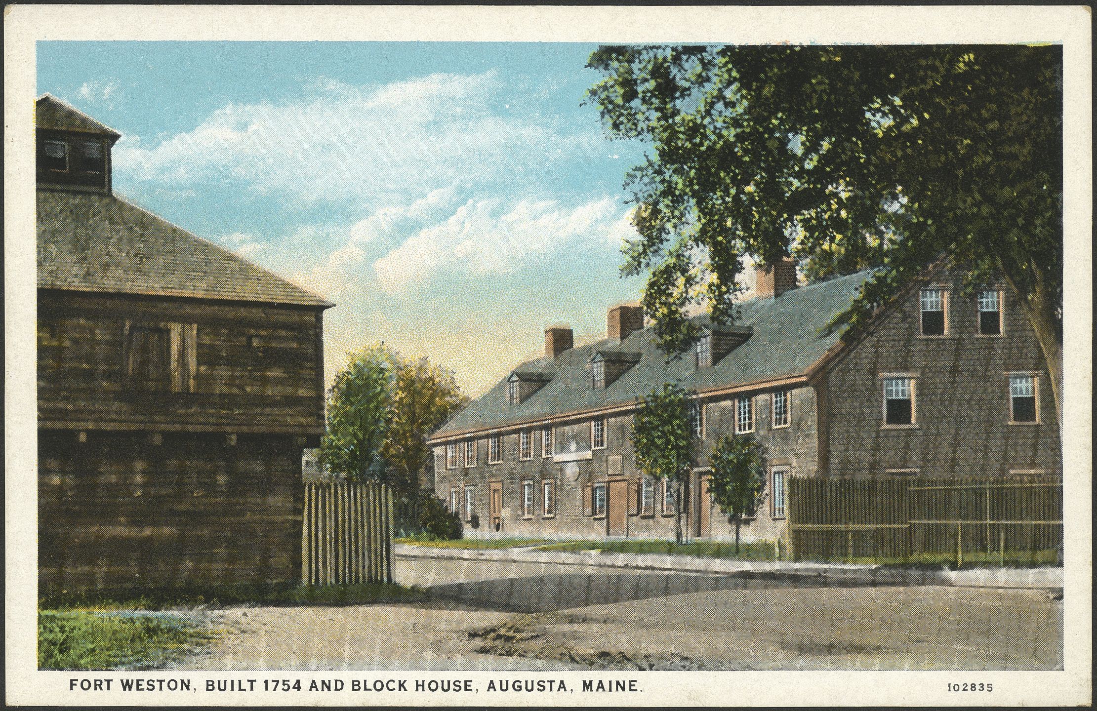 A vintage color postcard showing a rustic log blockhouse in the left foreground and a long, two-story wooden garrison building stretching along a road on the right under a blue sky.