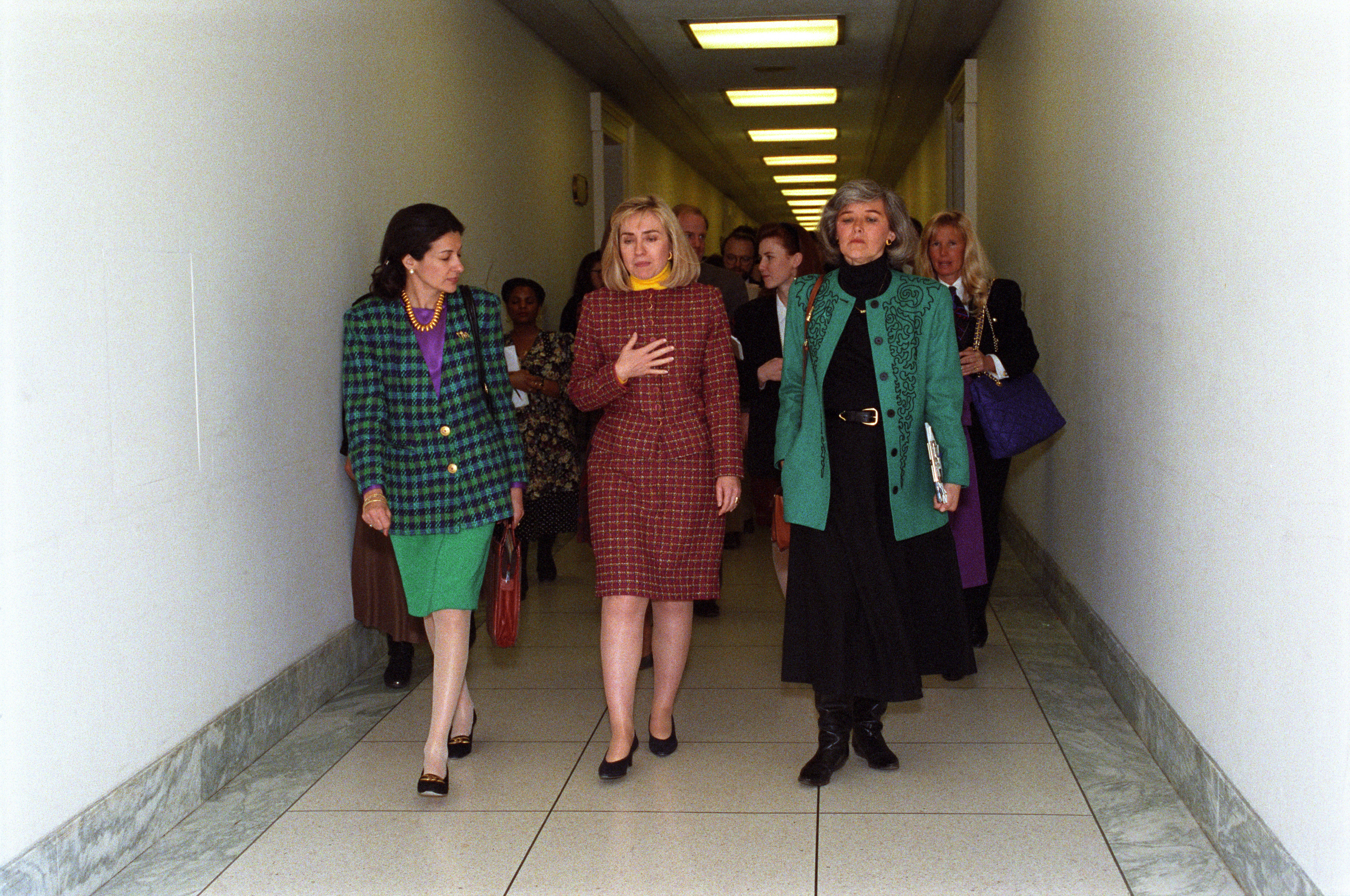 Olympia Snowe, wearing a green and blue plaid blazer, walks down a long hallway alongside First Lady Hillary Clinton, who is dressed in a red plaid skirt suit. They are followed by a small group of people under bright overhead fluorescent lights.