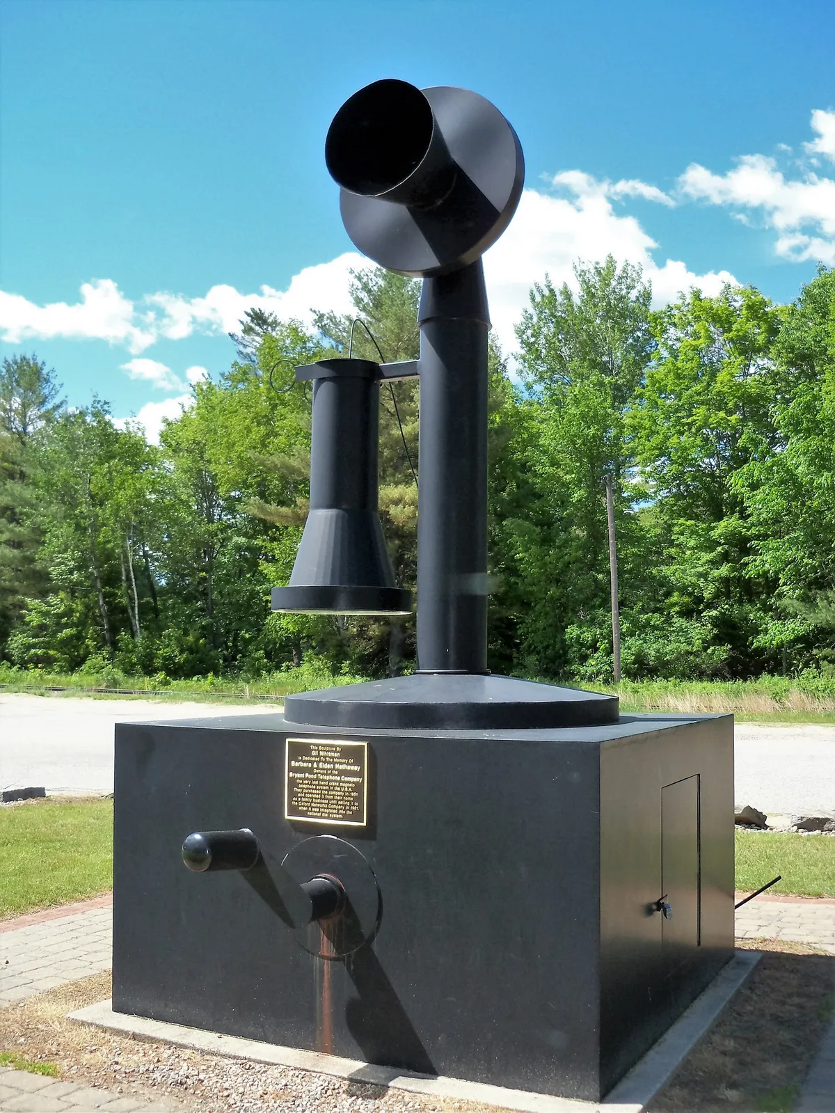 Outdoor sculpture of an oversized black candlestick telephone mounted on a granite pedestal, commemorating the Bryant Pond Telephone Company, the last hand-crank magneto telephone system in the U.S., with trees and blue sky behind.