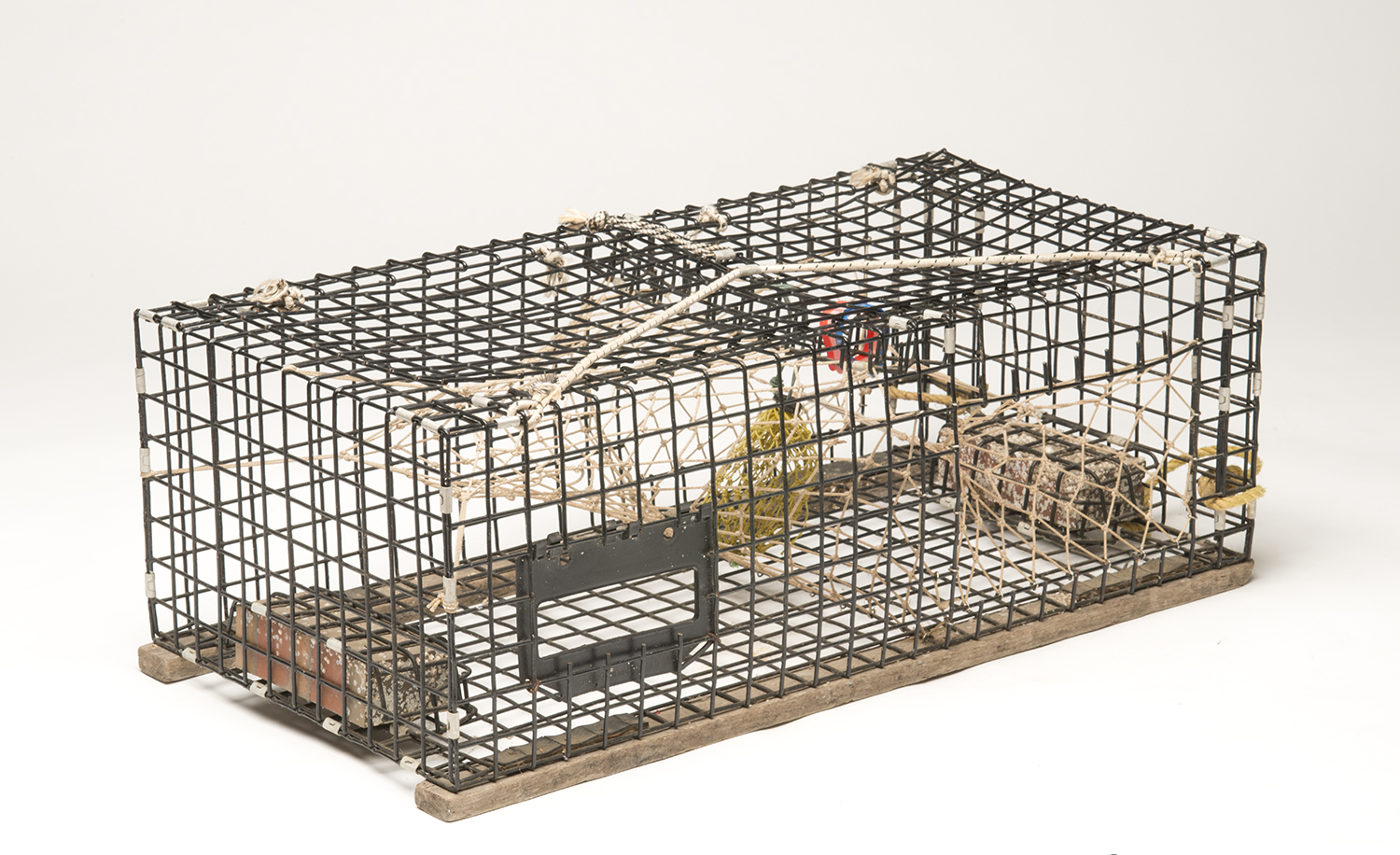 A studio product photograph of a modern rectangular lobster trap made of black vinyl-coated wire mesh, resting on a white background. Inside, the mesh netting funnels, bait bag, and bricks used for weight are clearly visible.