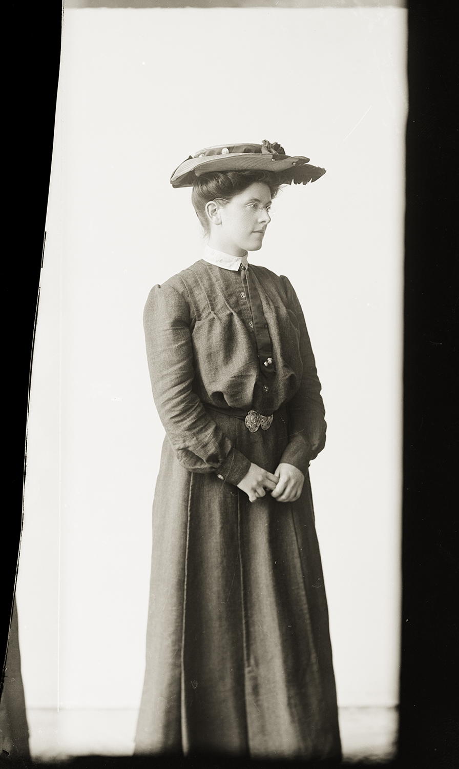 A black and white studio portrait of a woman captured from a three-quarters angle, standing against a plain white background. She is wearing an elegant, long-sleeved dark dress with a white collar, an ornate belt buckle, and a decorative wide hat perched on her styled hair.