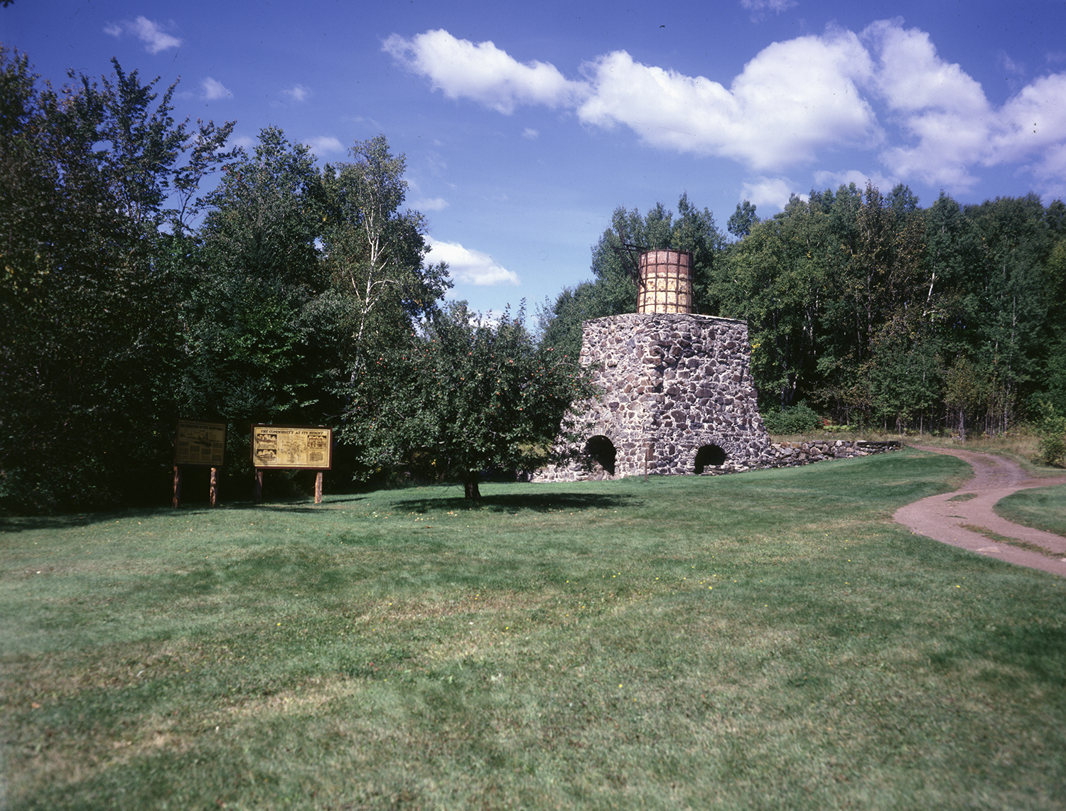 A vibrant color photograph of the historic stone blast furnace structure with its iron top stack, surrounded by lush green lawns, trees, informational signs, and a dirt path under a bright blue sky with fluffy white clouds.