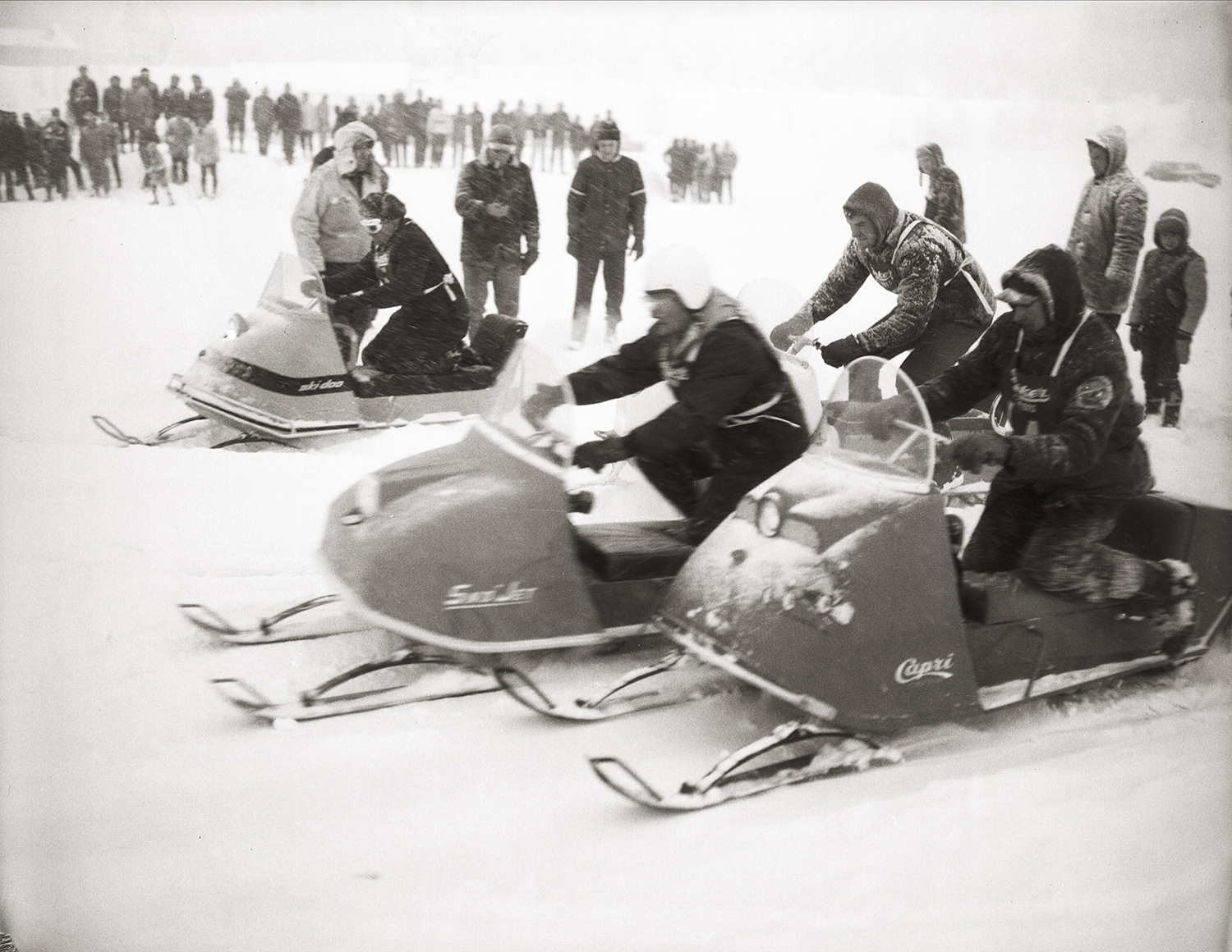 A historic, black and white photograph capturing action during a snowmobile race. Several vintage snowmobiles are driving across the snow, with a crowd of spectators bundled in winter clothing watching from the background.