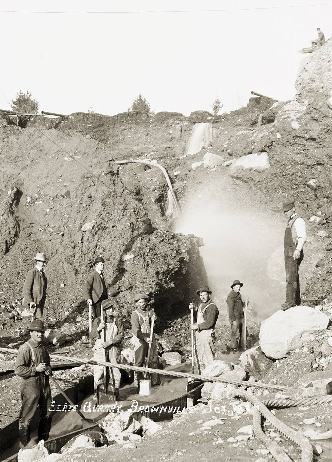 An archival sepia photograph showing a group of quarry workers posing amidst a rugged, muddy landscape with large rocks. Several men hold shovels near a wooden trough or flume, while others look toward the camera, and a white caption at the bottom reads "Slate Quarry, Brownville Jct., Me."