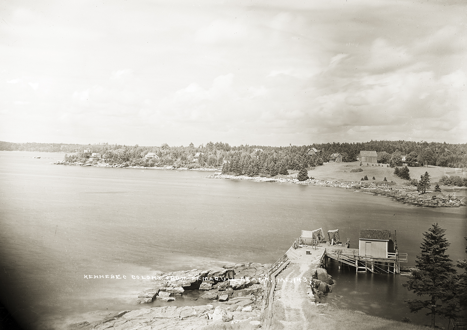 A black and white scenic landscape photograph overlooking a bay with a wooden pier and a small building in the lower right foreground. Wooded coastlines and scattered houses line the shore under a cloudy sky.