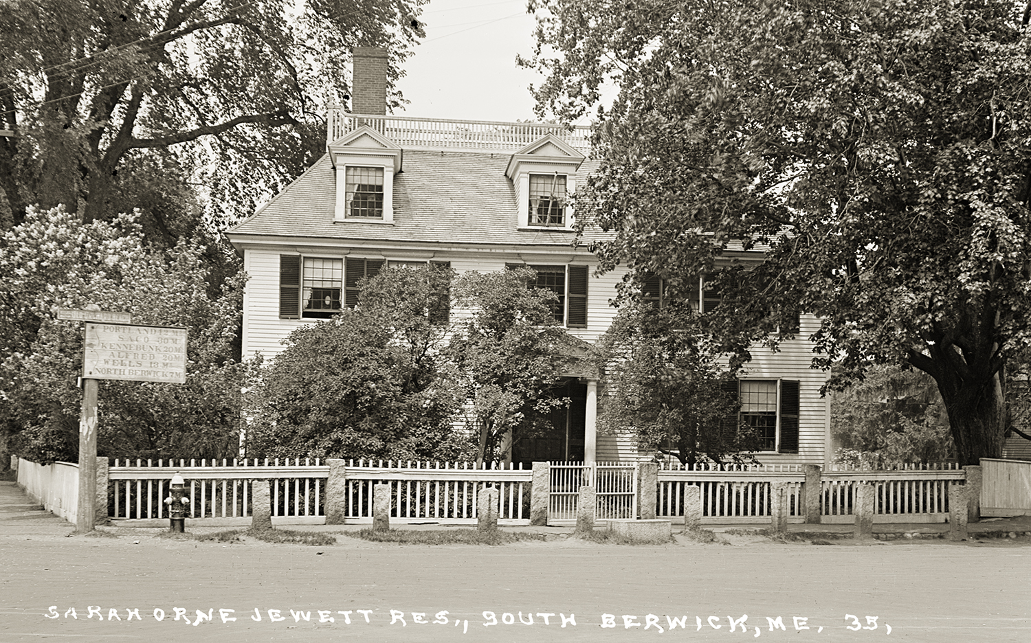 A vintage sepia-toned postcard showing the front view of the Sarah Orne Jewett Residence in South Berwick, Maine. Lush green bushes obscure the lower half of the two-story house behind a white fence, and a wooden signpost on the left lists mileage to nearby towns.