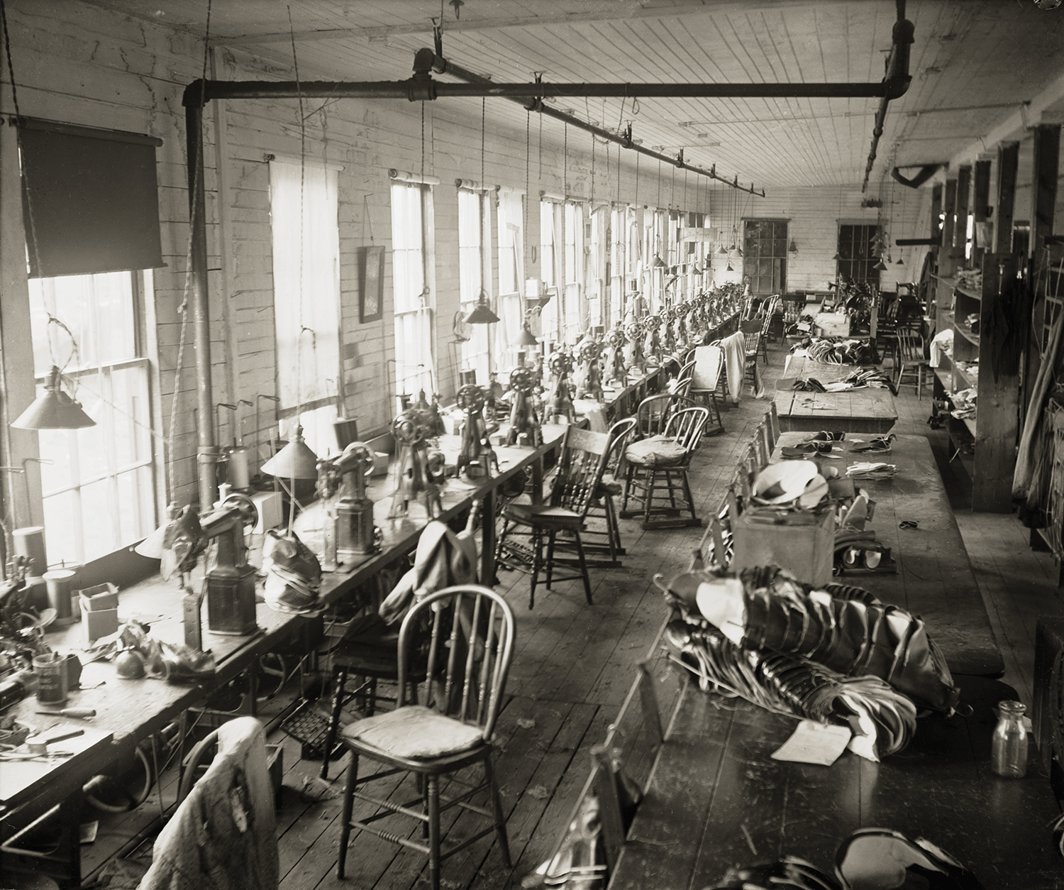 A black and white interior photograph of a long, narrow industrial workshop or stitching room. Multiple rows of vintage sewing machines line a long workbench on the left under hanging light fixtures, while wooden chairs sit empty and stacks of shoe components rest on tables to the right.