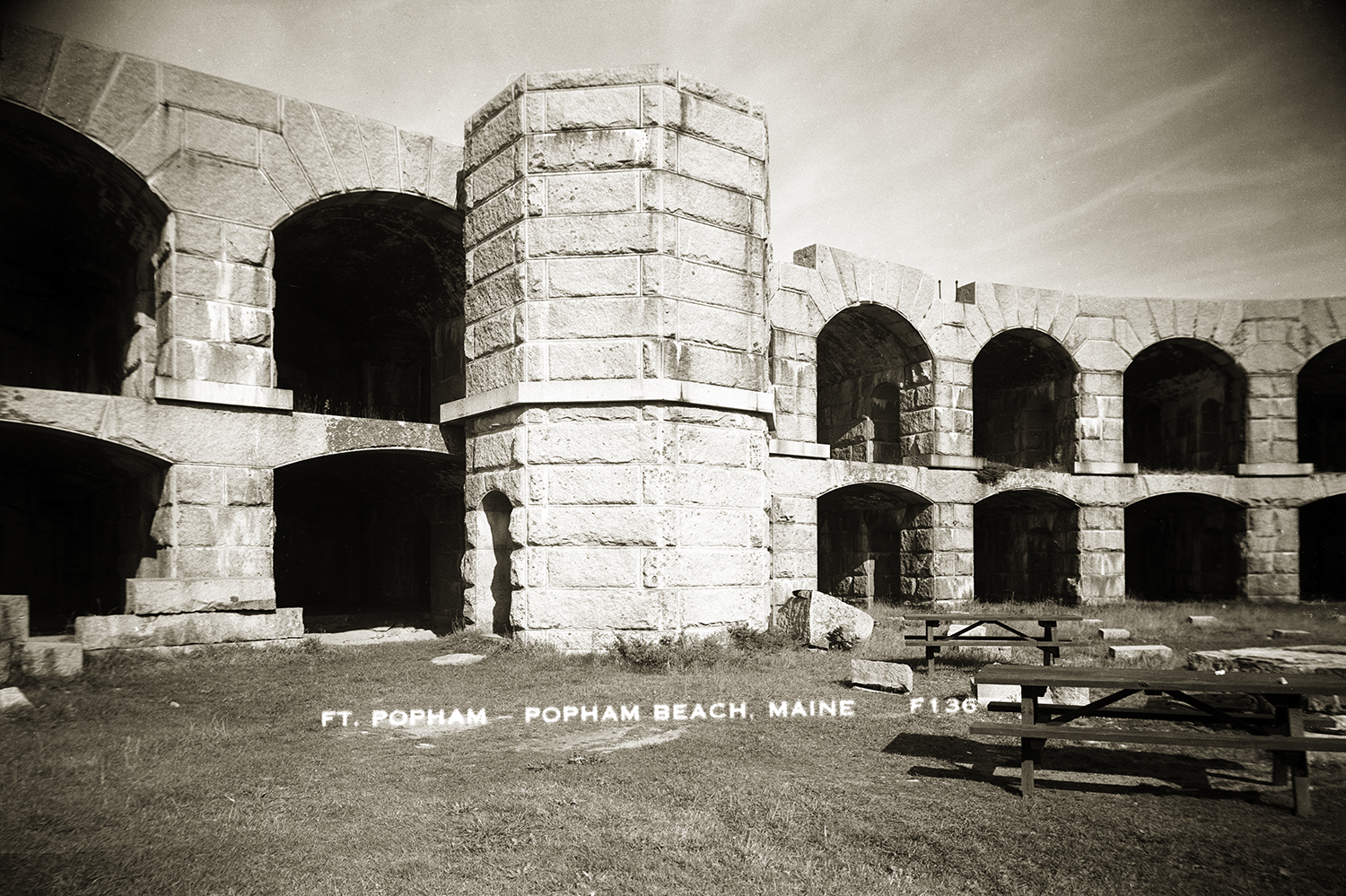 A sepia-toned vintage photograph showing the interior courtyard of Fort Popham in Popham Beach, Maine. The curved granite block structure features two tiers of open stone arches, with a central octagonal stone tower and two wooden picnic tables placed on the grass in the foreground.