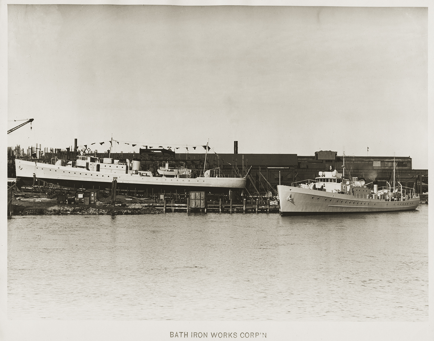 A black and white photograph capturing two military vessels at a shipyard facility, one resting on a dry dock slipway and the other afloat nearby, with a label at the bottom reading "BATH IRON WORKS CORP'N".