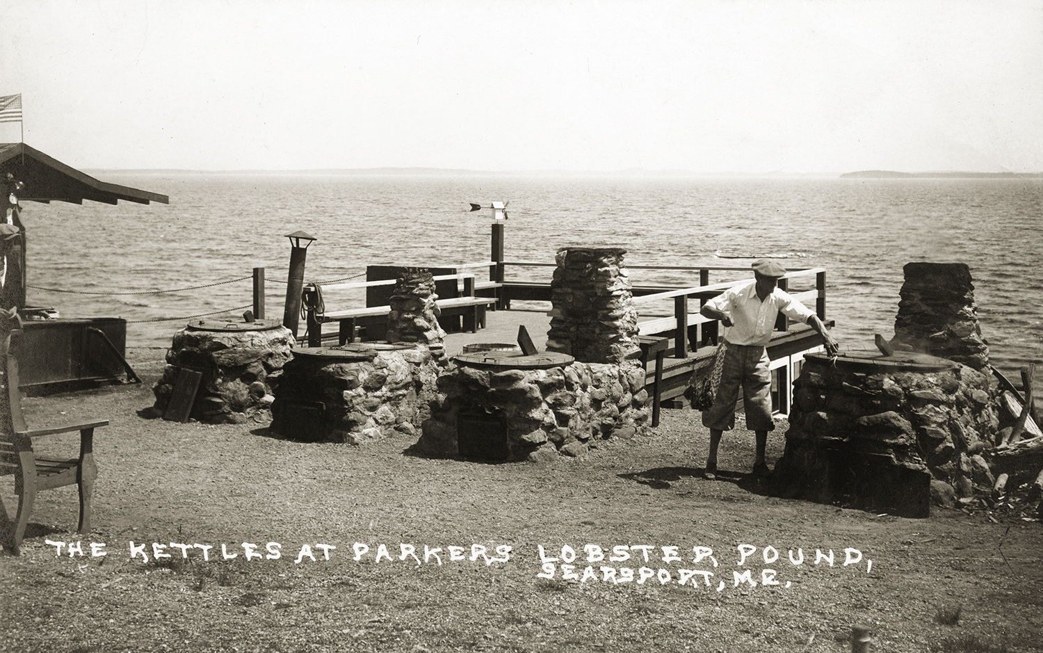 A vintage black and white photograph featuring a row of large stone and brick outdoor cooking kettles at an oceanfront property. A man stands near one of the smoking structures, and text at the bottom reads, "THE KETTLES AT PARKERS LOBSTER POUND, SEARSPORT, ME."