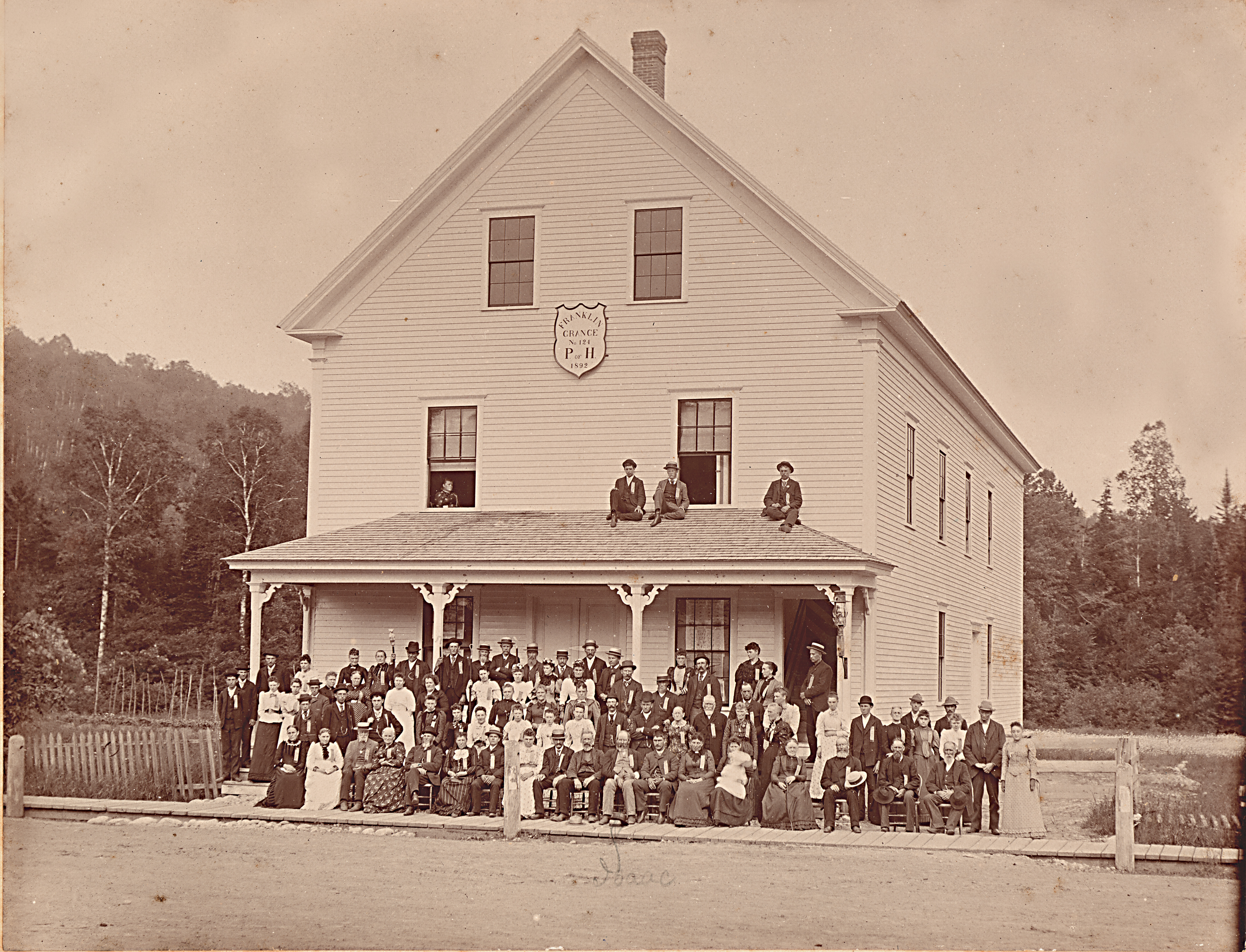 Large group of Grange members posed in front of the Franklin Grange No. 124 hall, with several young men sitting on the porch roof and a shield reading "Franklin Grange No. 124, P. of H., 1892."