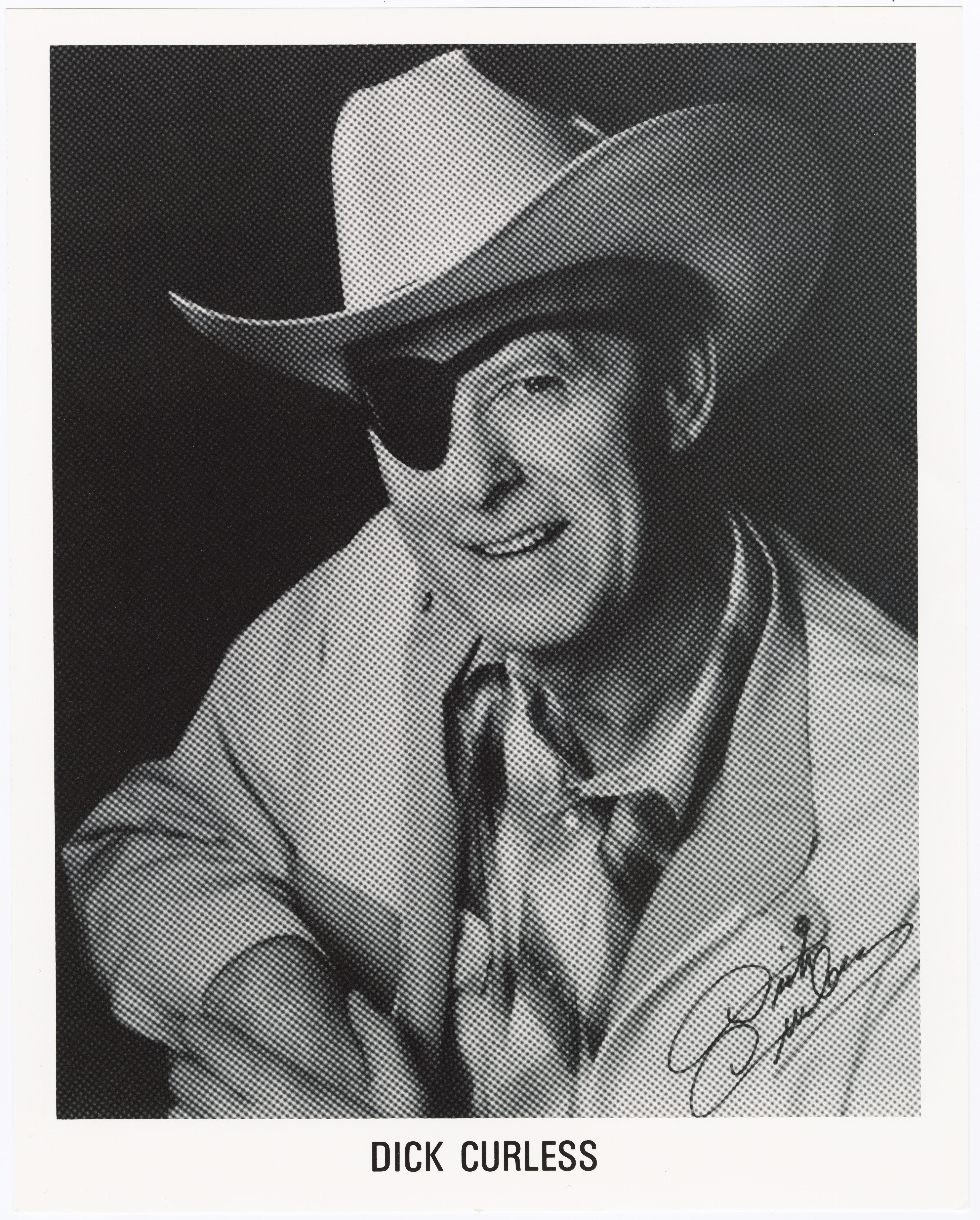 Black-and-white publicity portrait of country singer Dick Curless wearing a white cowboy hat and eye patch, smiling and signed with his autograph in the lower right corner.